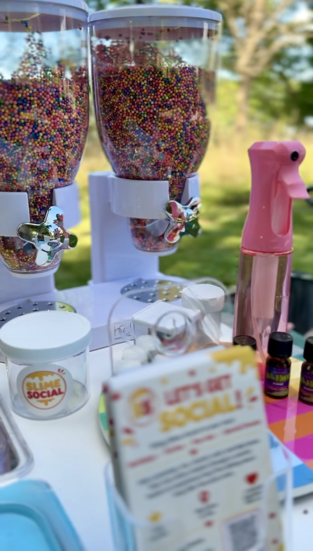 Two clear containers filled with colorful tiny candy sprinkles, with a pink and clear spray bottle nearby, and various small items on a white surface outdoors.