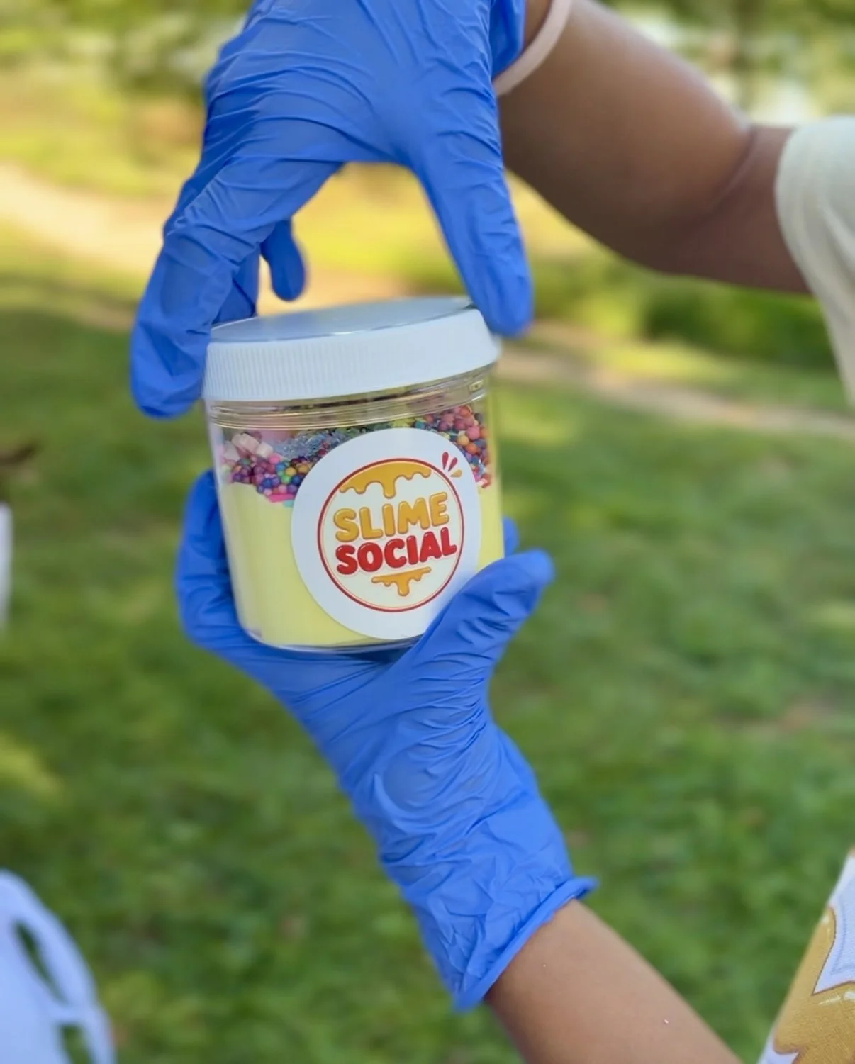 A person wearing blue gloves holding a clear jar titled 'Slime Social' with colorful slime inside.