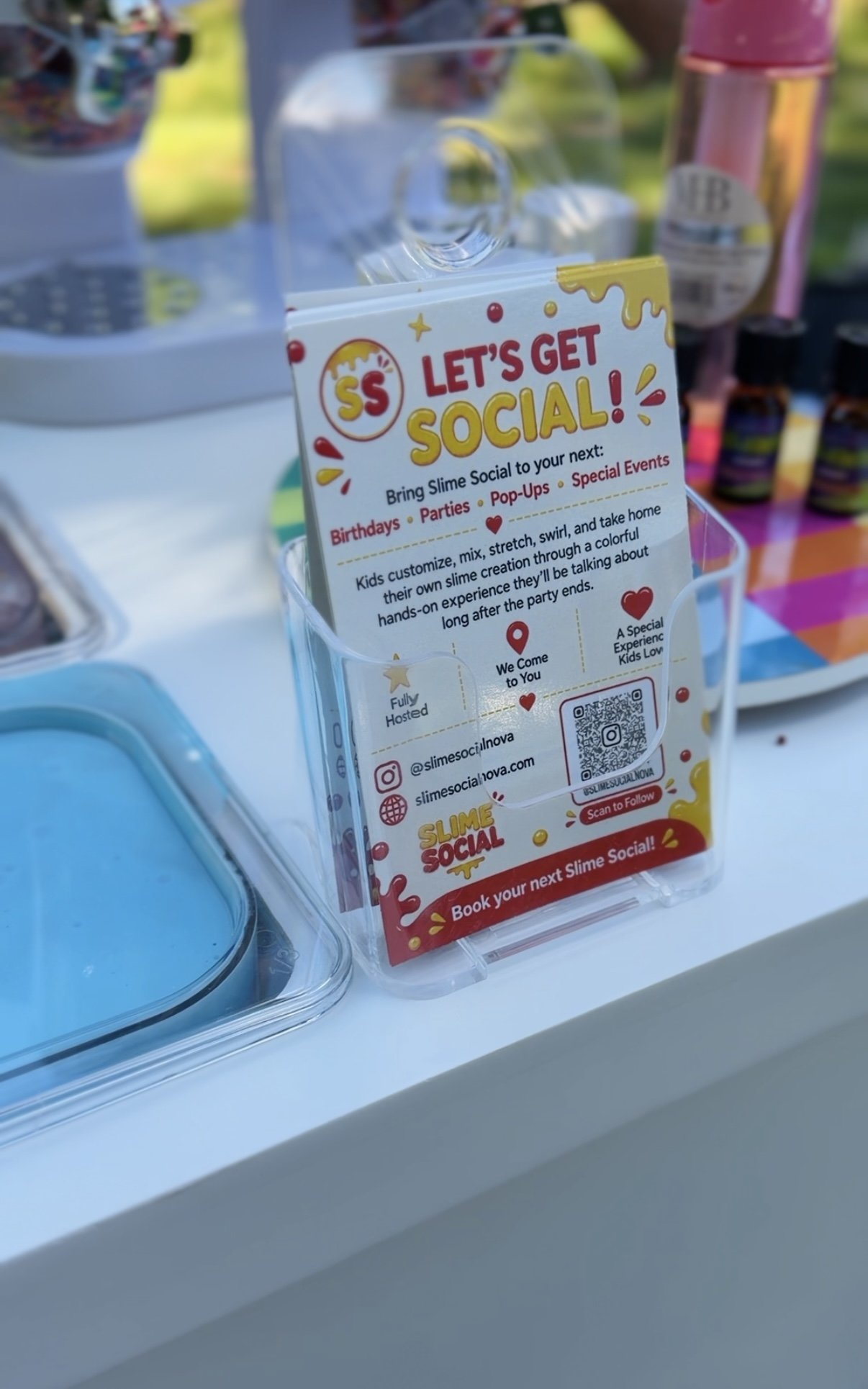 A colorful sign promoting slime social events such as birthdays, parties, pop-ups, and special events. The sign invites kids to customize, mix, stretch, swirl, and take home their slime creation. It includes social media handles, a QR code, and a message to book the next slime social event.
