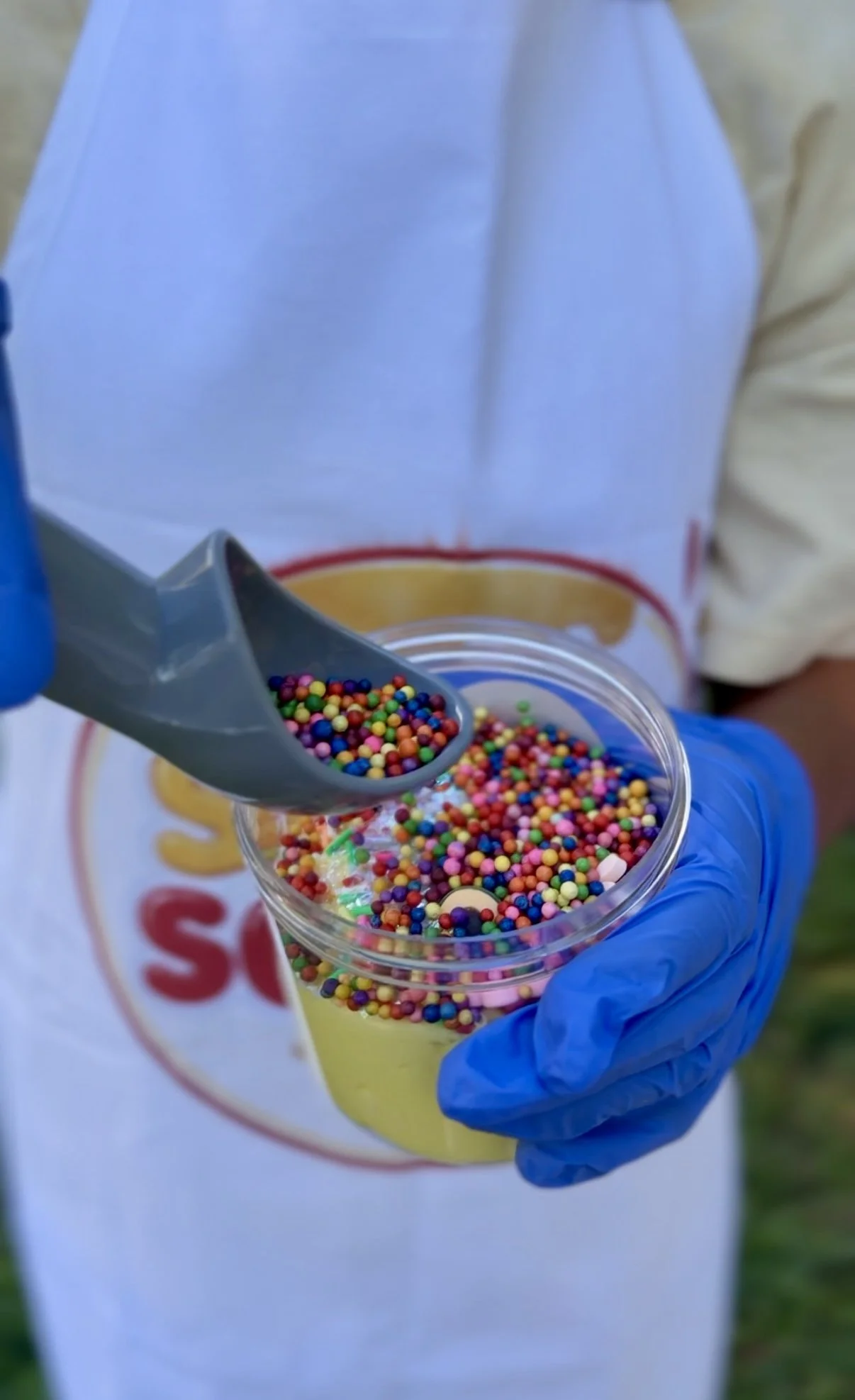 Person wearing blue gloves scooping colorful sprinkles from a container into a cup.