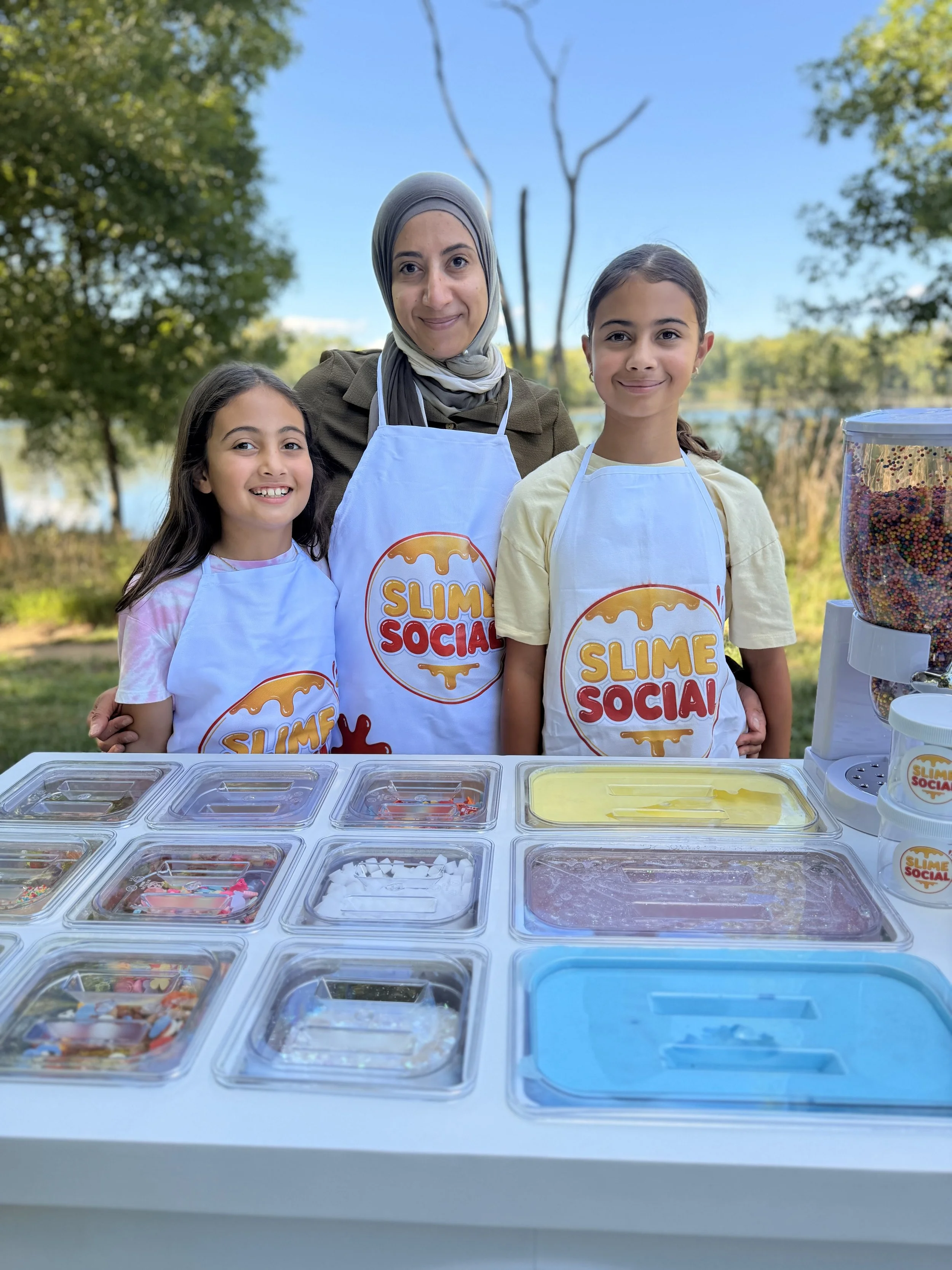 Three girls and a woman smiling behind a table of colorful slime jars outdoors during daytime. They are wearing aprons with a slime logo, and the background features trees and a lake.
