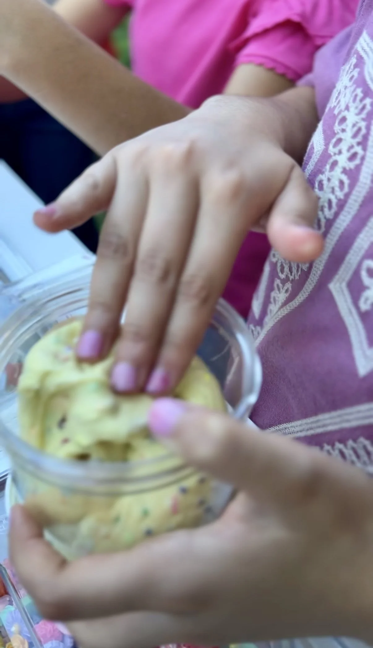 Close-up of child's hand reaching into a jar of yellow cookies or crackers, with other children in colorful clothing visible in the background.