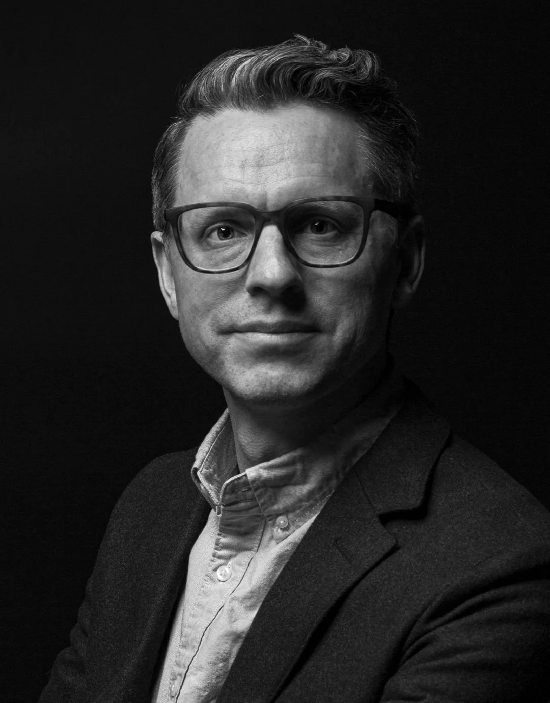 Black and white headshot portrait of Philip Cheaney. He is wearing glasses, a collared shirt and blazer, seated against a dark background.