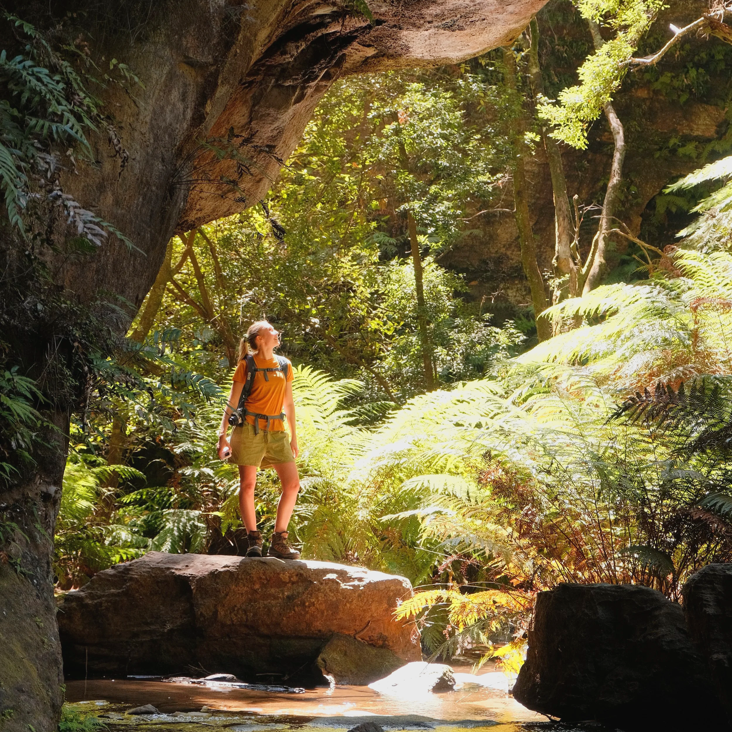 A woman wearing an orange shirt, beige shorts, and hiking boots stands on a rock in front of a small waterfall in a lush, green forest.