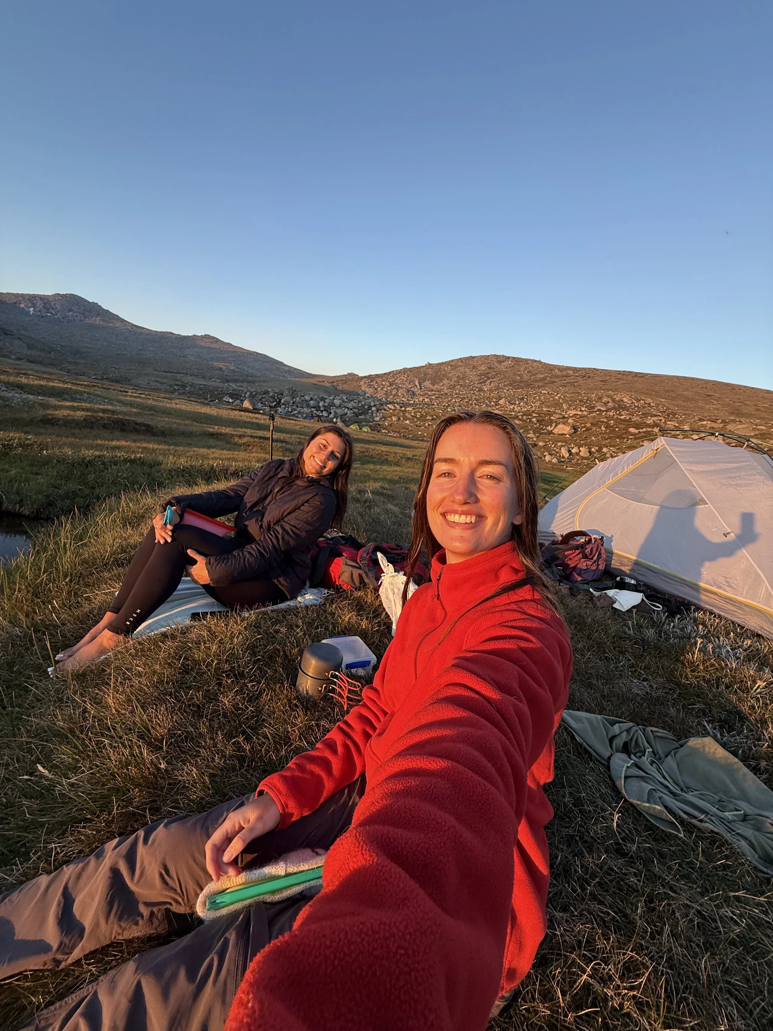 Two women camping outdoors in a grassy field with mountains in the background during sunset. One woman is taking a selfie, and both are smiling. There is a tent and camping gear around them.