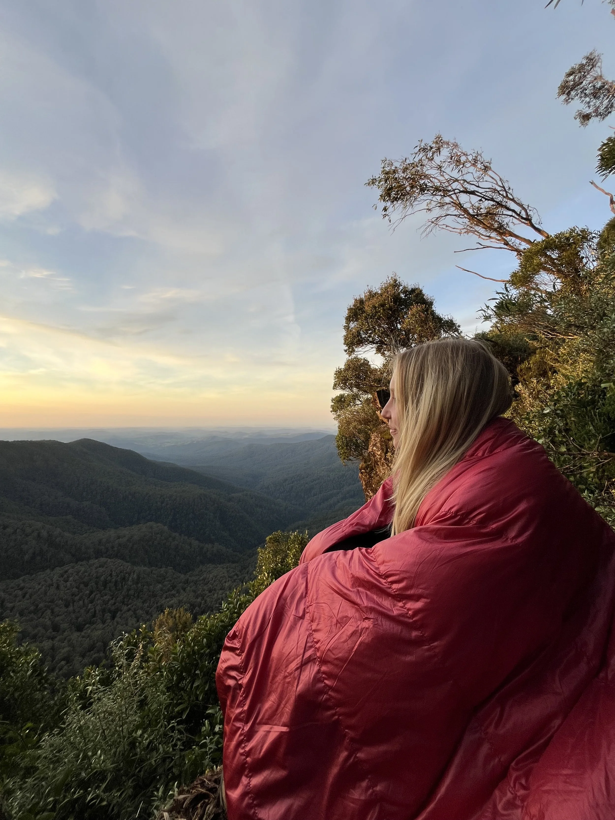 Person with blonde hair wearing sunglasses and a red jacket, sitting on a hillside overlooking a mountain view during sunset.
