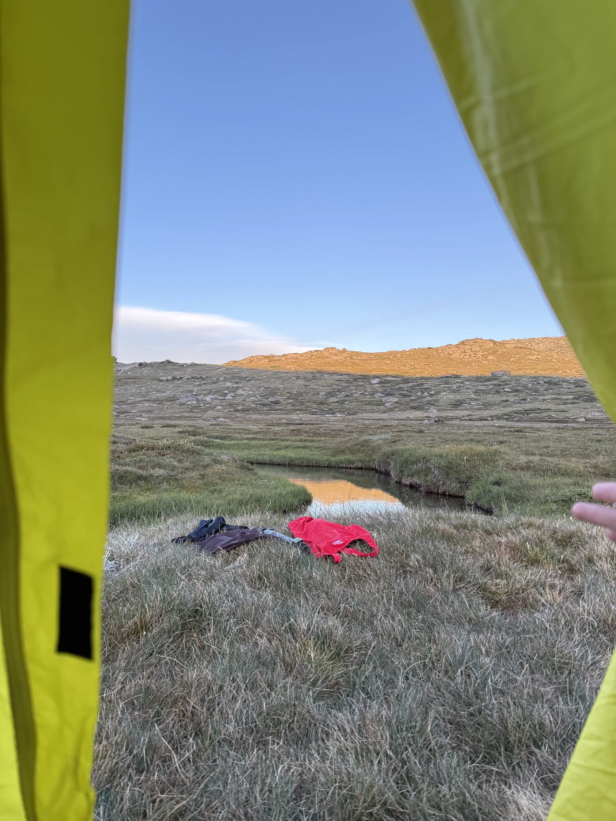 View of a landscape with a small stream, grassy terrain, hills or cliffs in the distance, visible through a gap in yellow fabric or material.