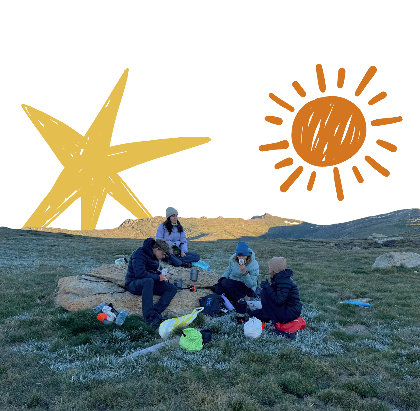 Four people sitting on a rocky patch in a grassy field with mountains in the background, enjoying an outdoor meal with camping gear and supplies around them.