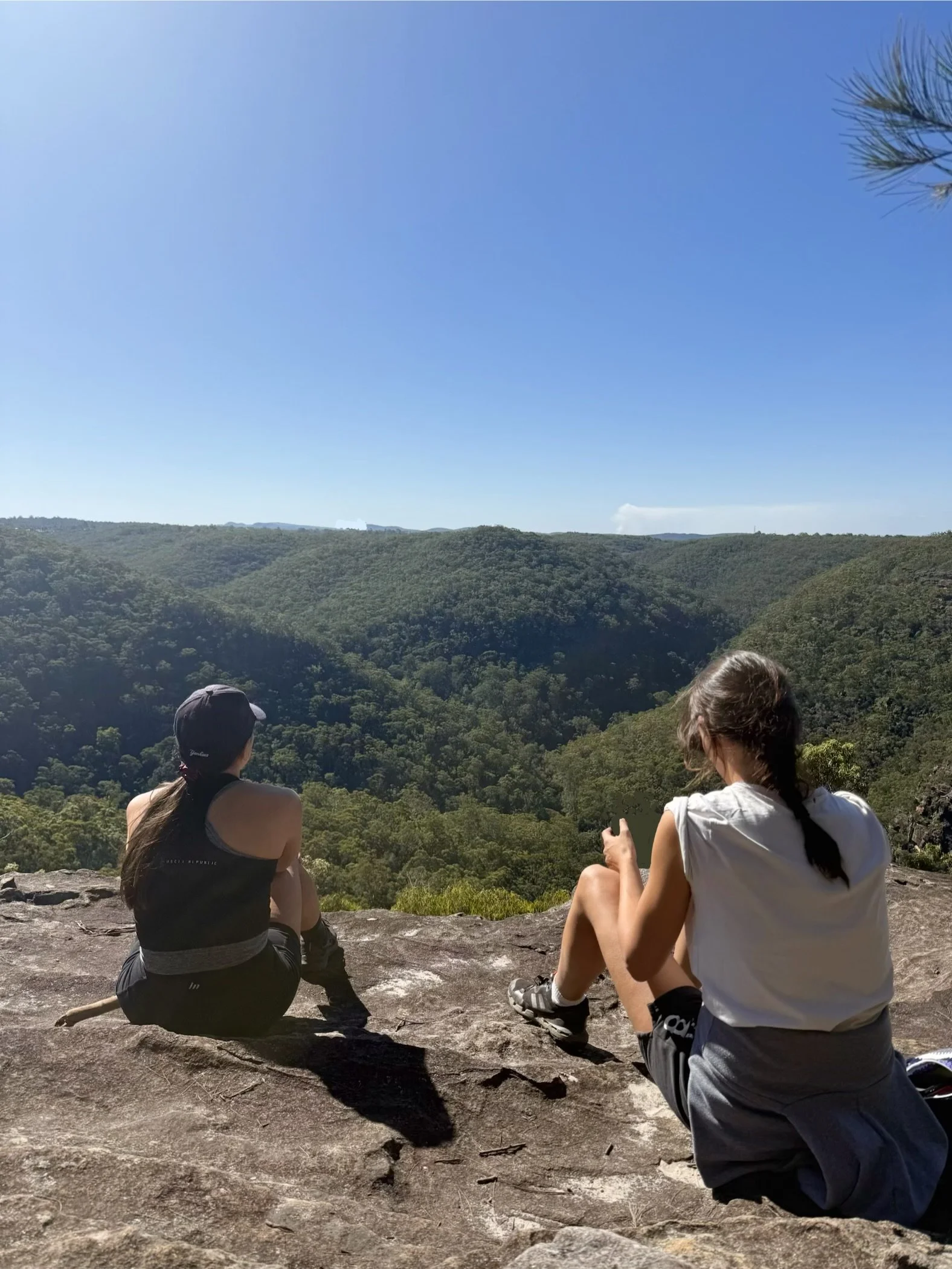 Two women sit on a rocky ledge overlooking a lush green landscape with rolling hills and a clear blue sky. One woman has long hair, a dark sports bra, and black shorts, while the other has her hair in a braid, a white shirt, and gray shorts. They are enjoying the outdoor view.