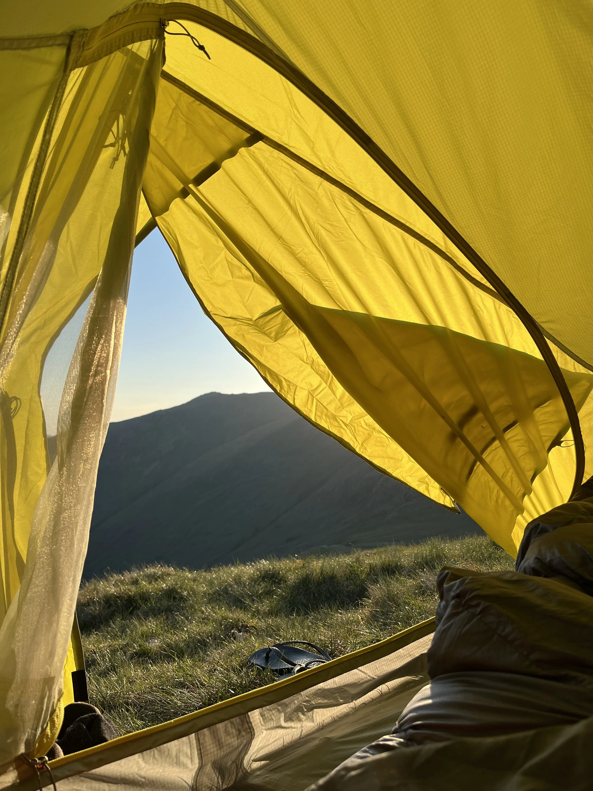 Inside a yellow tent with a mountain and blue sky in the background, daytime, with camping gear and grass on the ground.