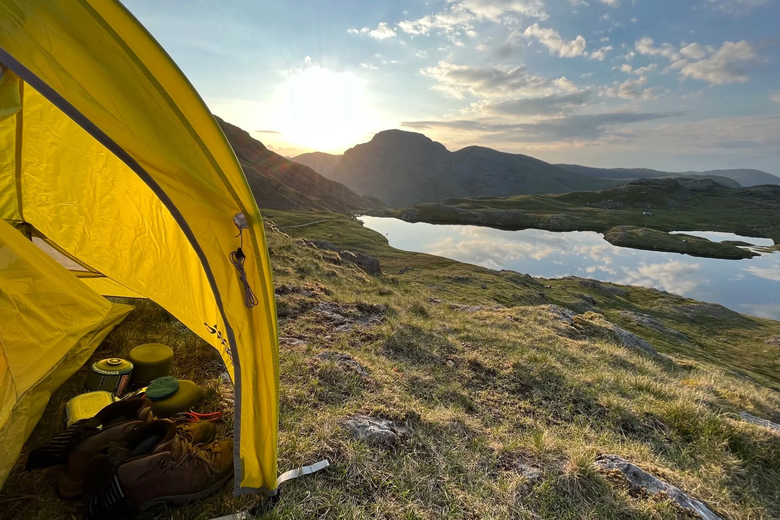 A yellow tent with shoes and gear outside, set in a mountainous landscape during sunset, with a body of water reflecting the sky and distant mountains.