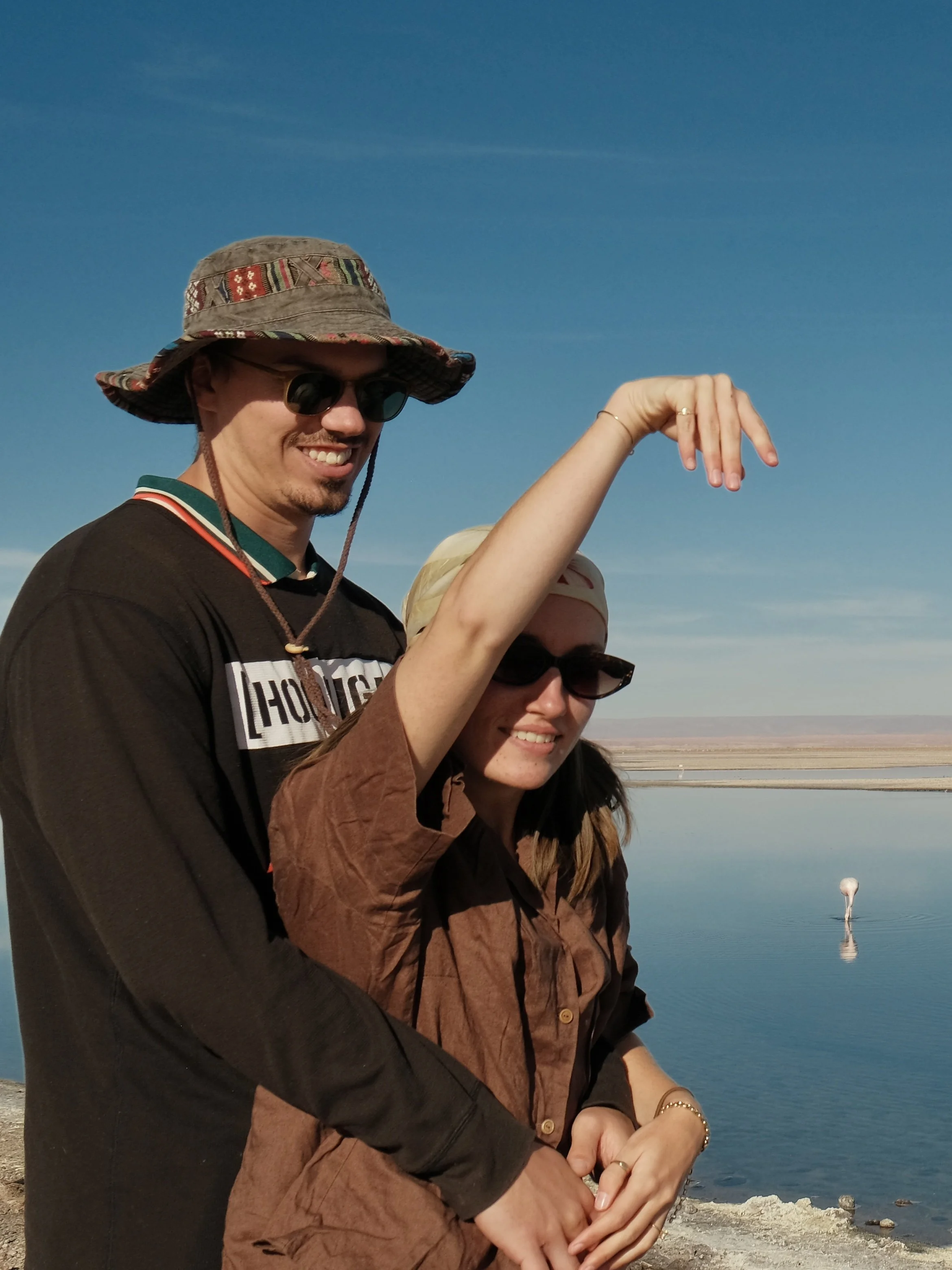 A smiling man in sunglasses and a wide-brimmed hat stands with a woman near a body of water under a clear blue sky, with a distant landmass or salt flat in the background.