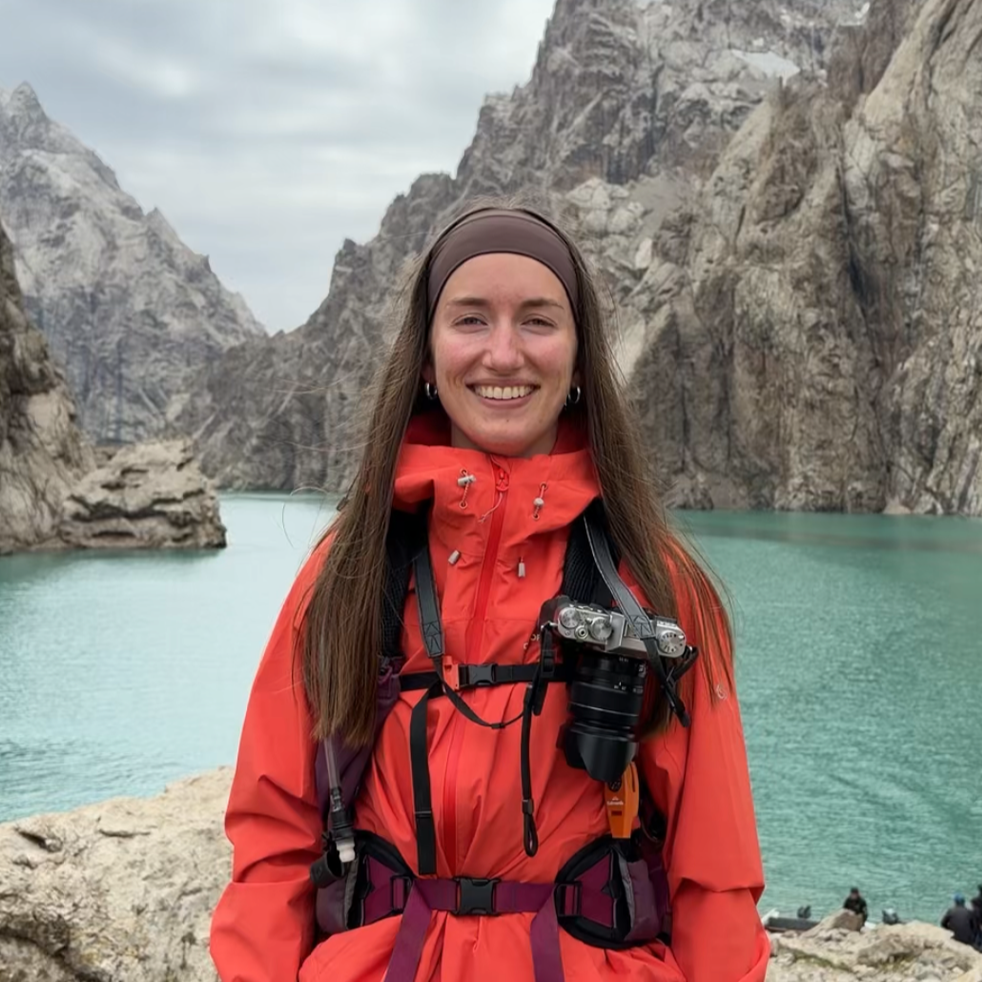 A woman in an orange jacket and headband smiling near a lake with rocky mountains in the background.