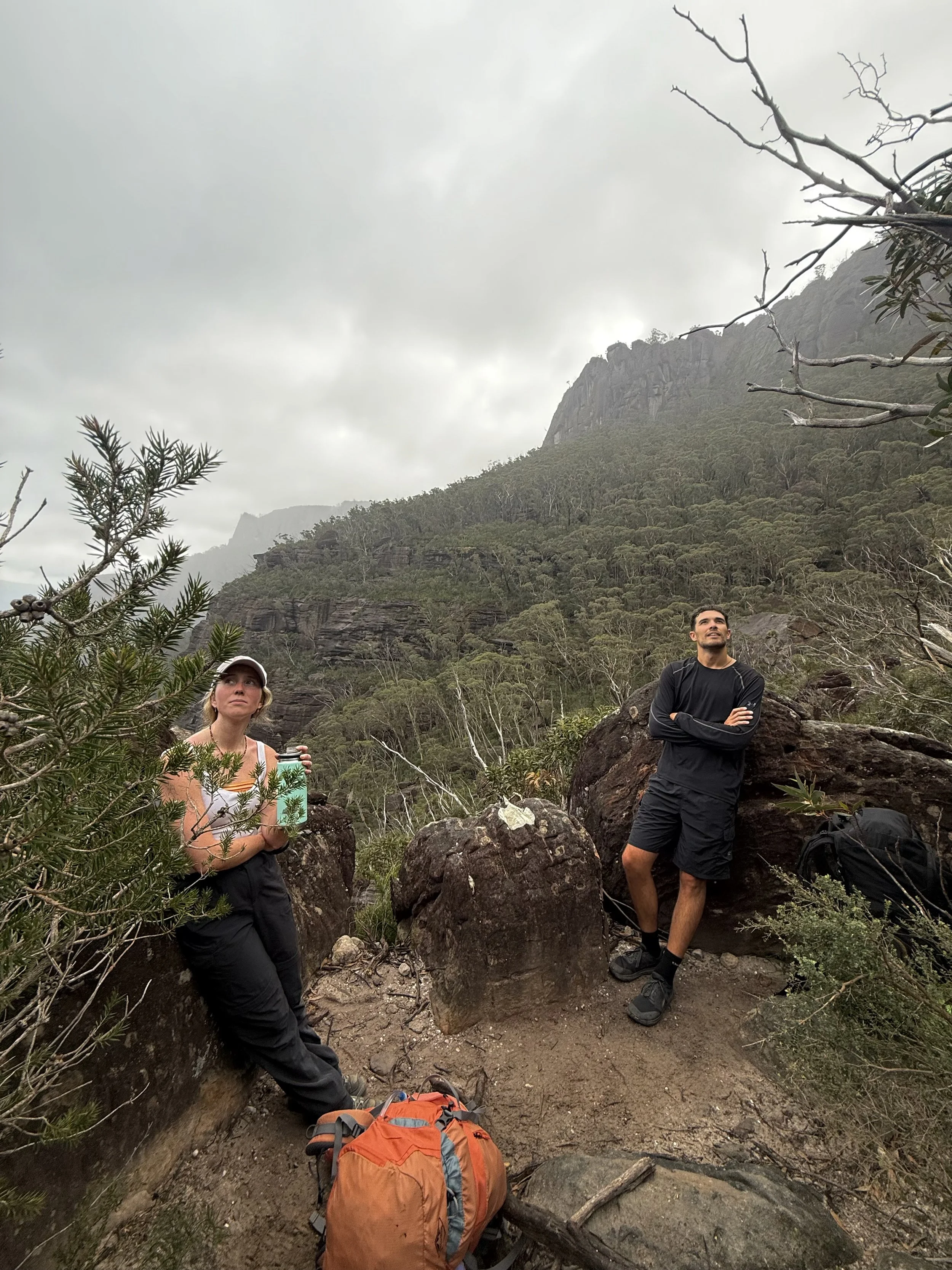 Two hikers on a guided overnight hike in Budawang National Park standing among rocks in a mountainous forested area under cloudy skies, with hiking gear and backpacks nearby.
