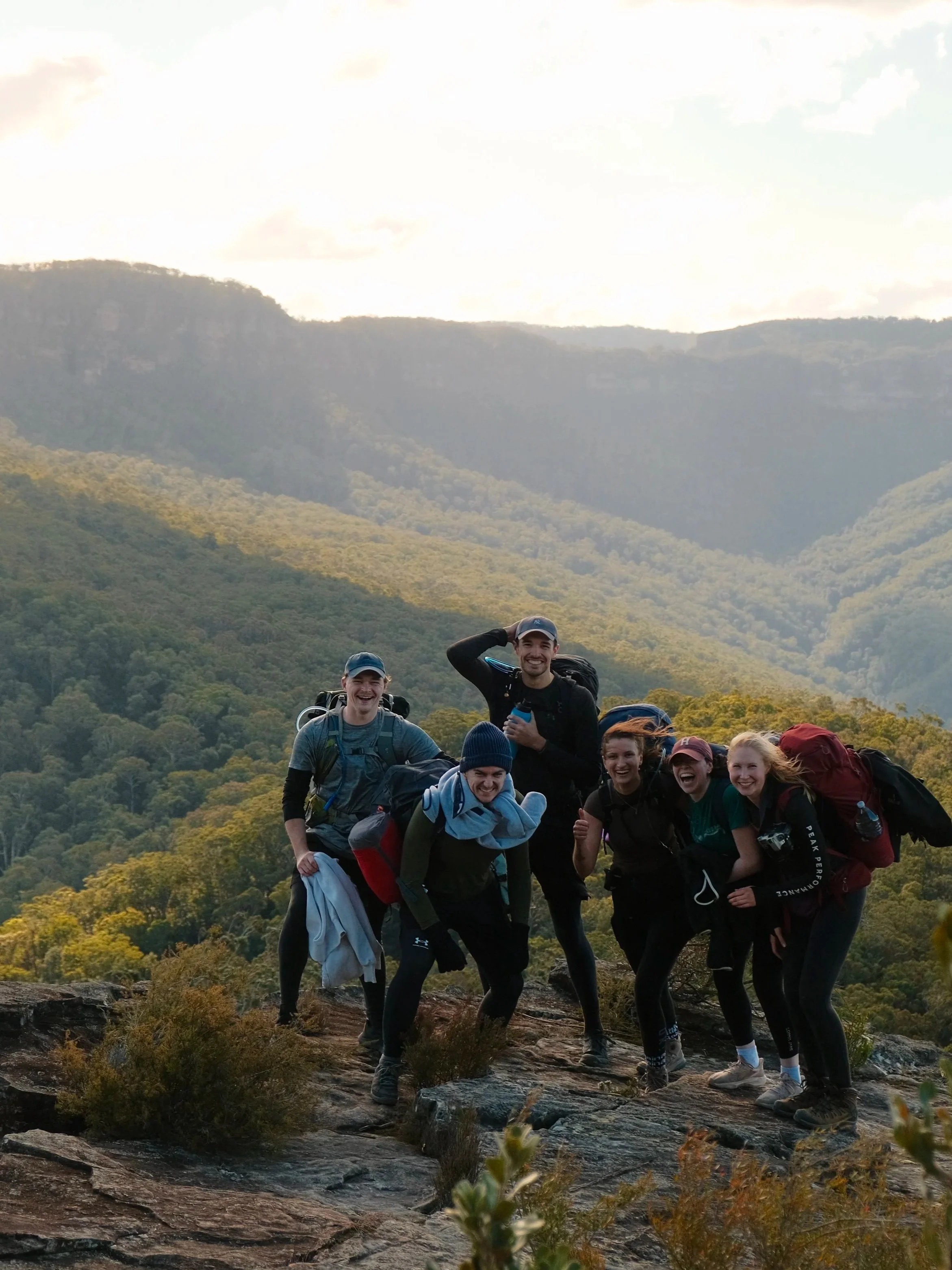 A group of seven young hikers with backpacks and outdoor clothing smiling and posing for a photo on a rocky trail with a mountain landscape and forest in the background. They are on a guided overnight hike in Blue Mountains National Park, in NSW