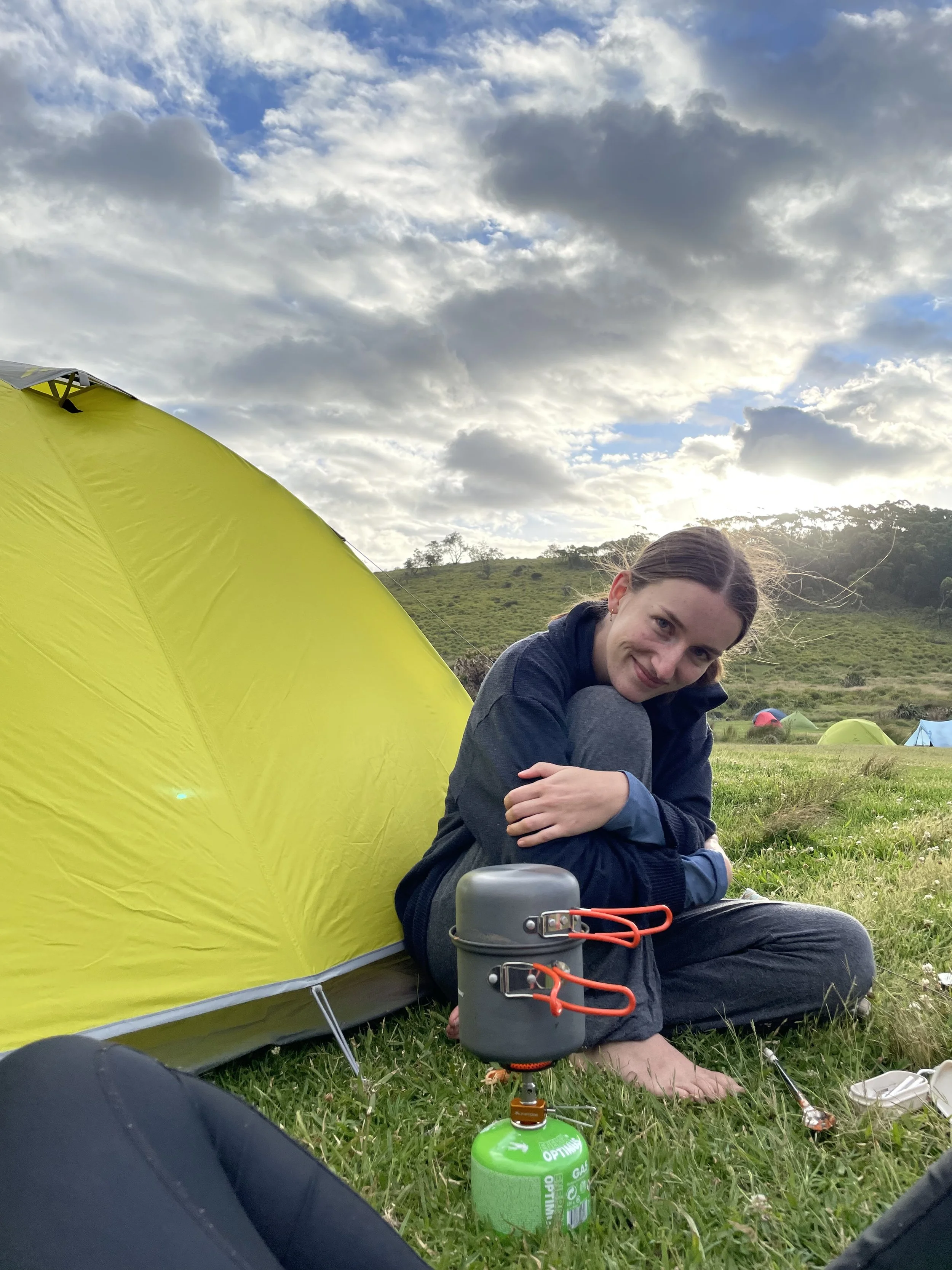 A woman sitting on grass next to a yellow tent, smiling at the camera, with camping gear and a green portable stove nearby in an outdoor grassy area with hills and cloudy sky in the background.