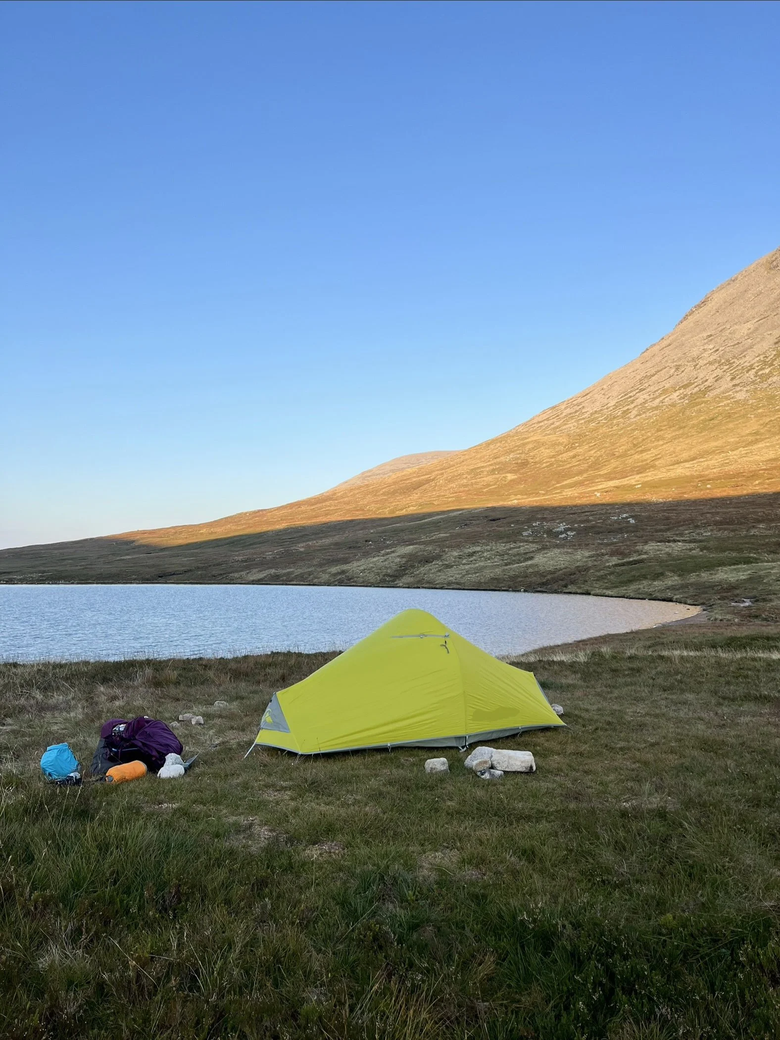 A yellow tent set up on grassy terrain by a lake, with mountains in the background, during daytime.