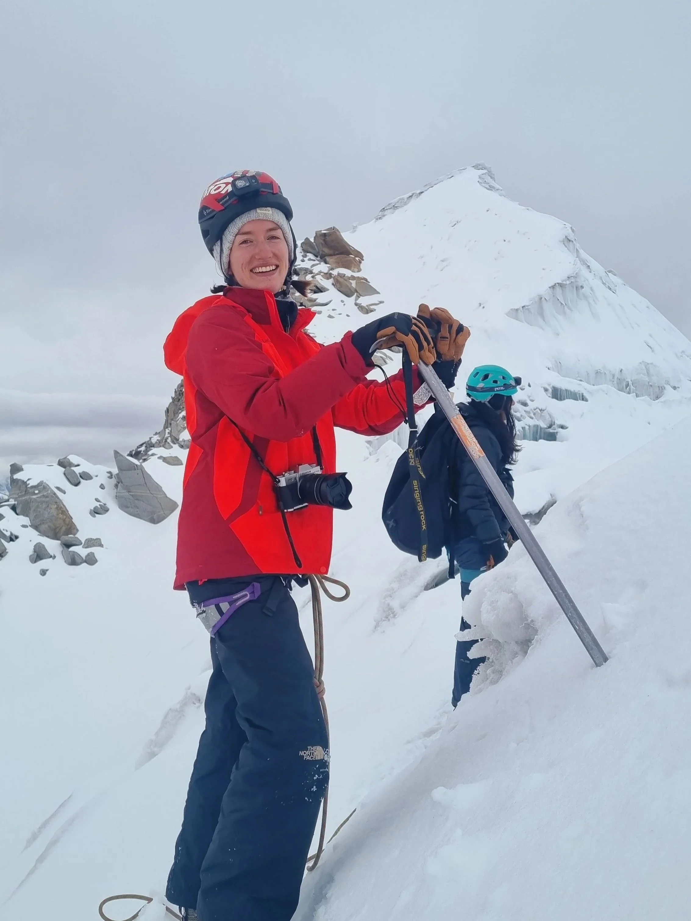 A woman climbing snowy mountain in winter gear, smiling, wearing a helmet, gloves, and carrying an ice axe, with snowy peaks and rocks in the background.