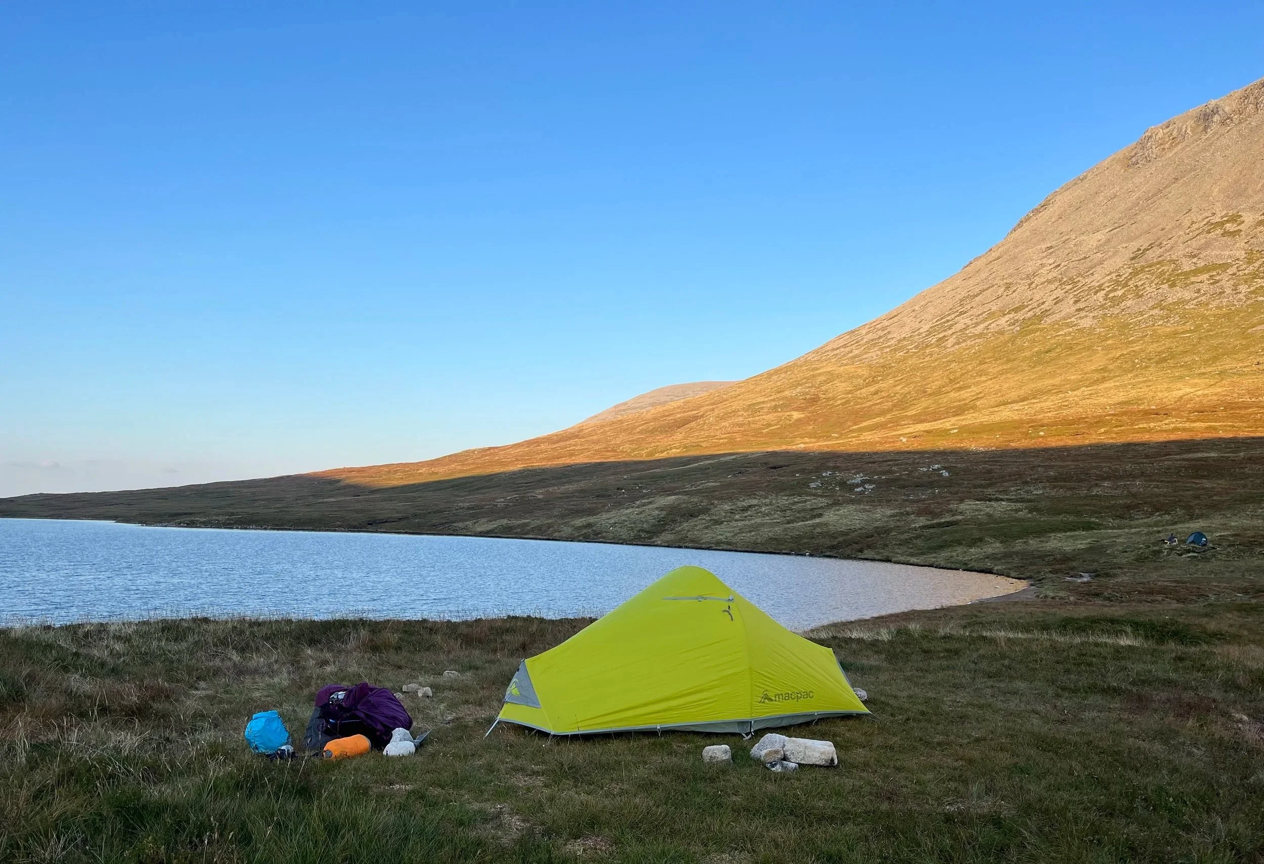 A yellow tent and camping gear set up on a grassy lakeshore, with a mountain in the background and a clear blue sky.