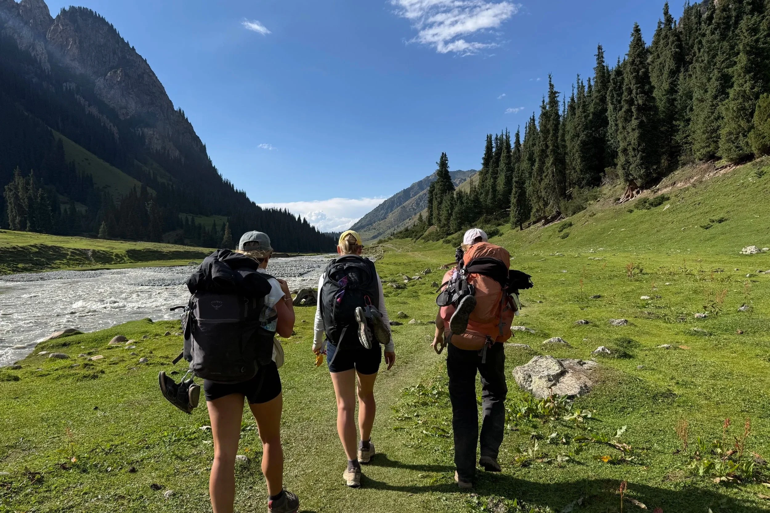 Group of hikers walking along a river in a green valley surrounded by tall mountains and dense trees under a partly cloudy sky.