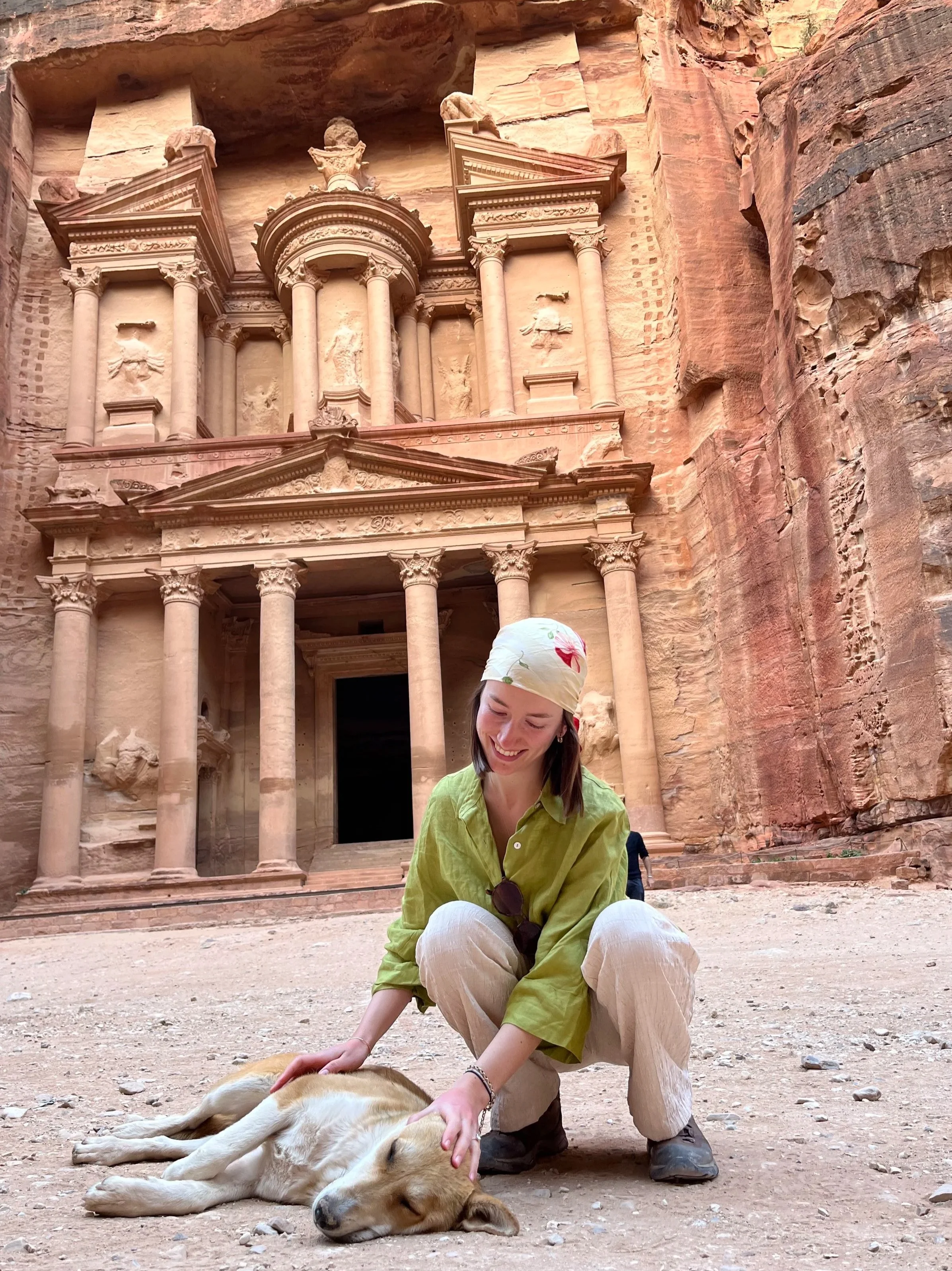 A woman in a green shirt and beige pants is petting a large, resting dog lying on the ground. They are outside near the ancient Al-Khazneh (The Treasury) carved into the red sandstone cliffs at Petra, Jordan.