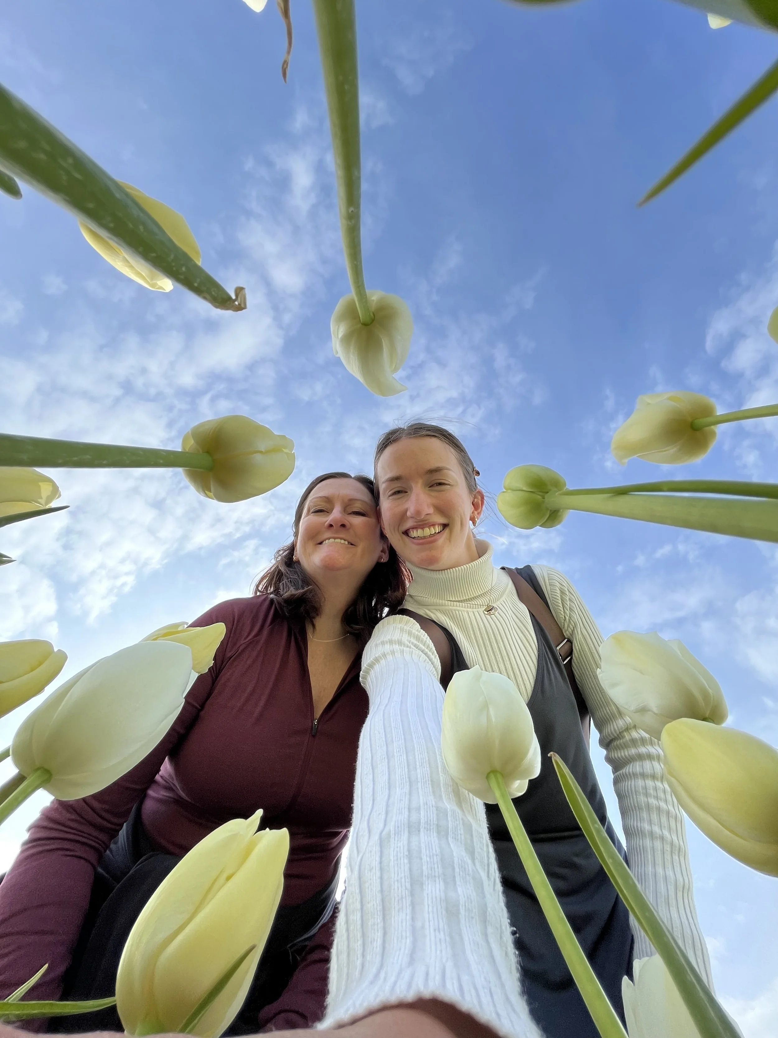 A selfie of two smiling women outdoors, taken from a ground-up perspective, surrounded by yellow tulips and a bright blue sky with some clouds.