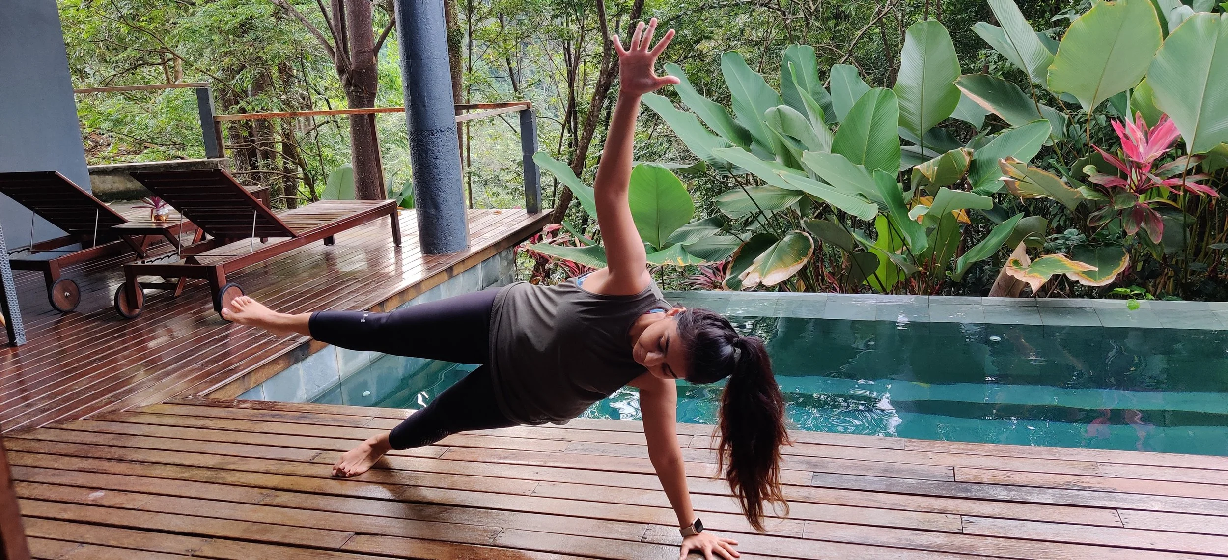 A woman doing a side plank yoga pose by a pool on a wooden deck, surrounded by lush tropical plants.