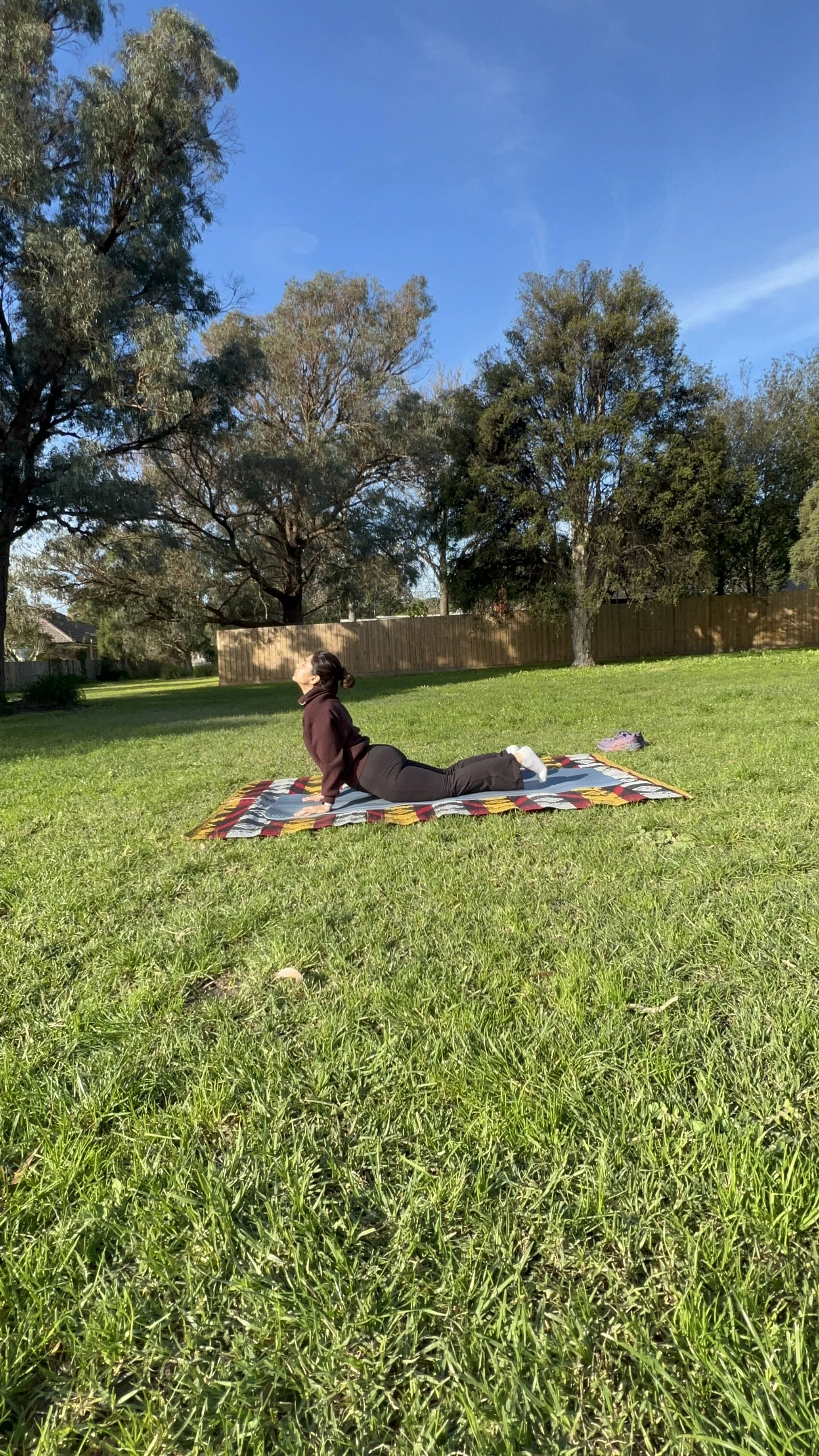 A woman practicing yoga outdoors on a sunny day, performing the Cobra pose on a colorful mat in a grassy yard with trees and a wooden fence in the background.