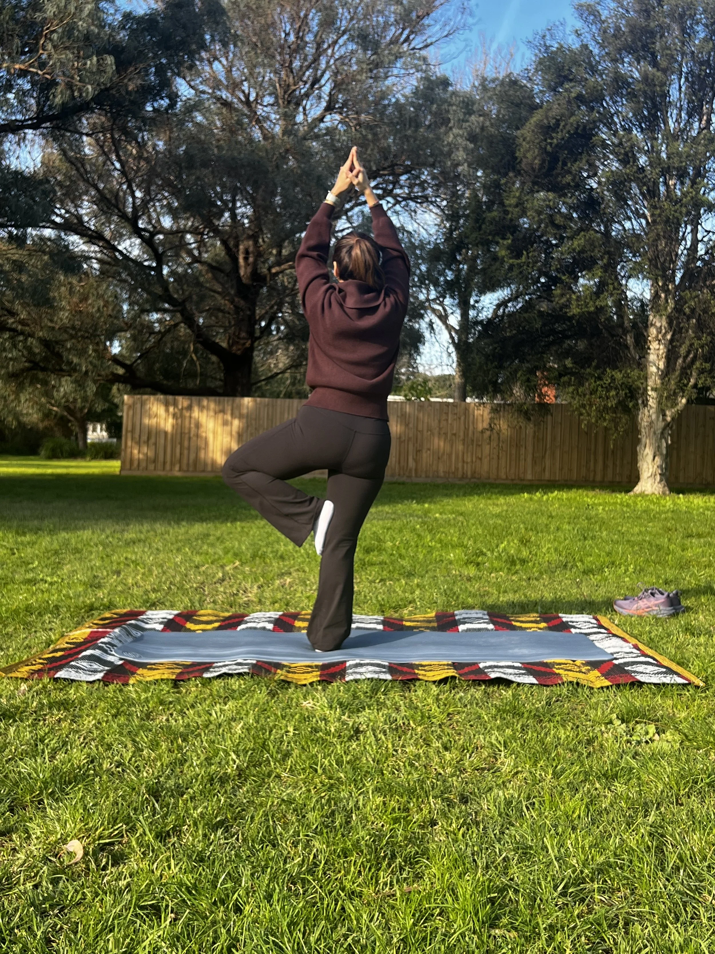 A woman practicing yoga outdoors on a blanket in a grassy yard with trees and a wooden fence in the background. She is in a tree pose with hands in prayer position overhead.