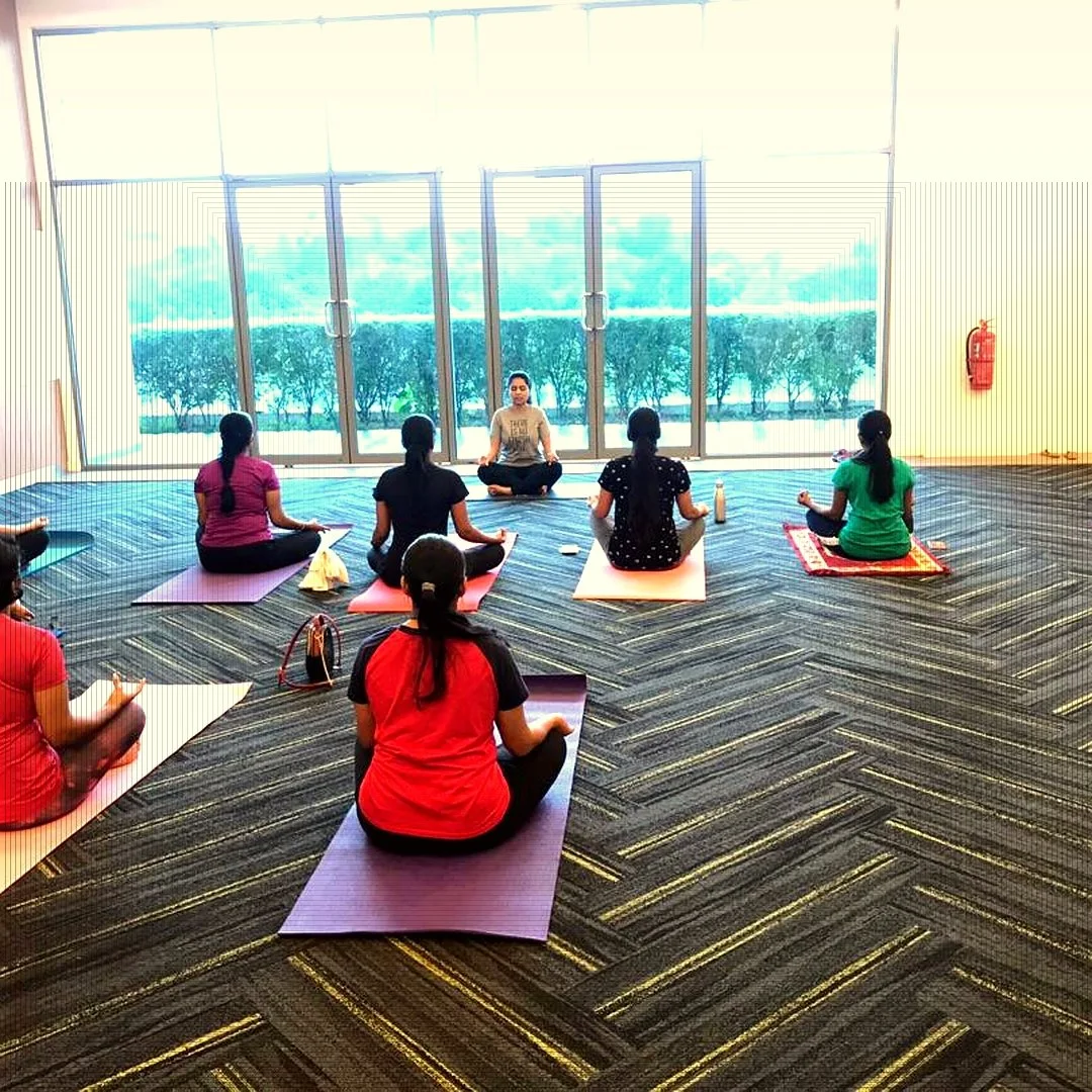 A group of women participating in a yoga class indoors on mats, seated in a cross-legged meditation pose. The instructor is leading the session towards a large glass window with a view of trees outside.