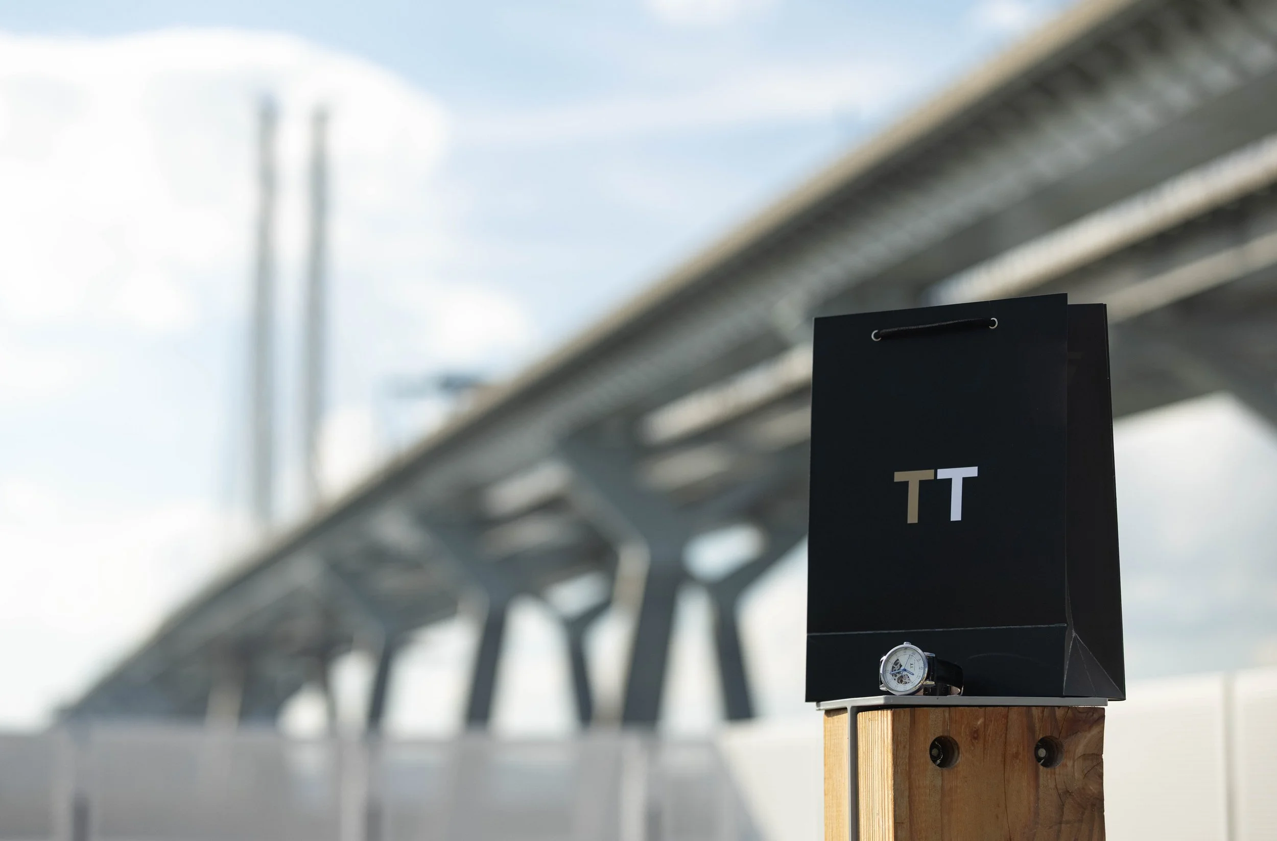 Close-up of a black sign with white text reading 'TT' attached to a wooden post, under a modern bridge structure with a clear sky background.