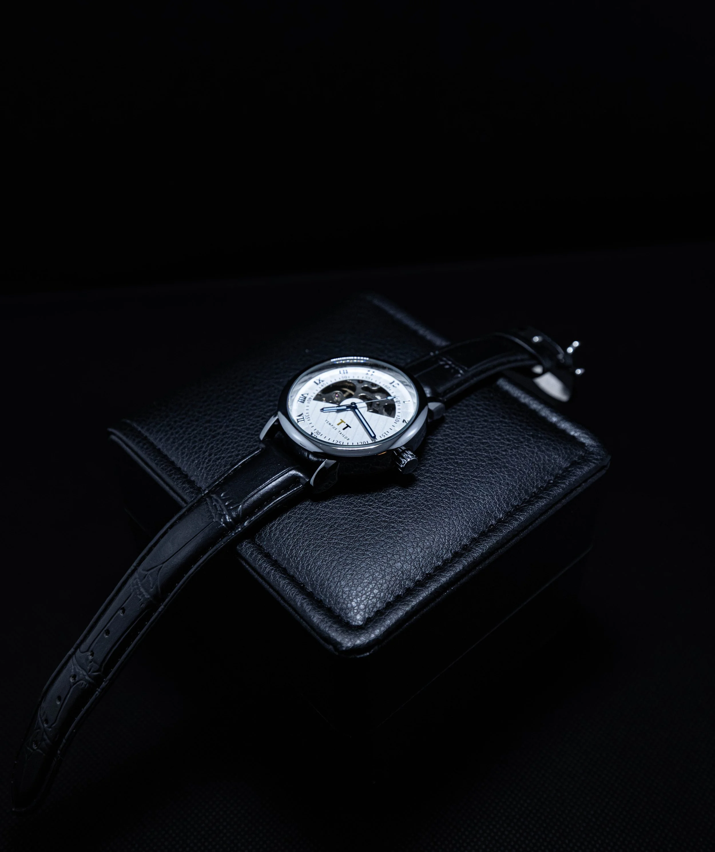 A wristwatch with a white face, black leather strap, and Roman numerals, resting on a black textured surface.