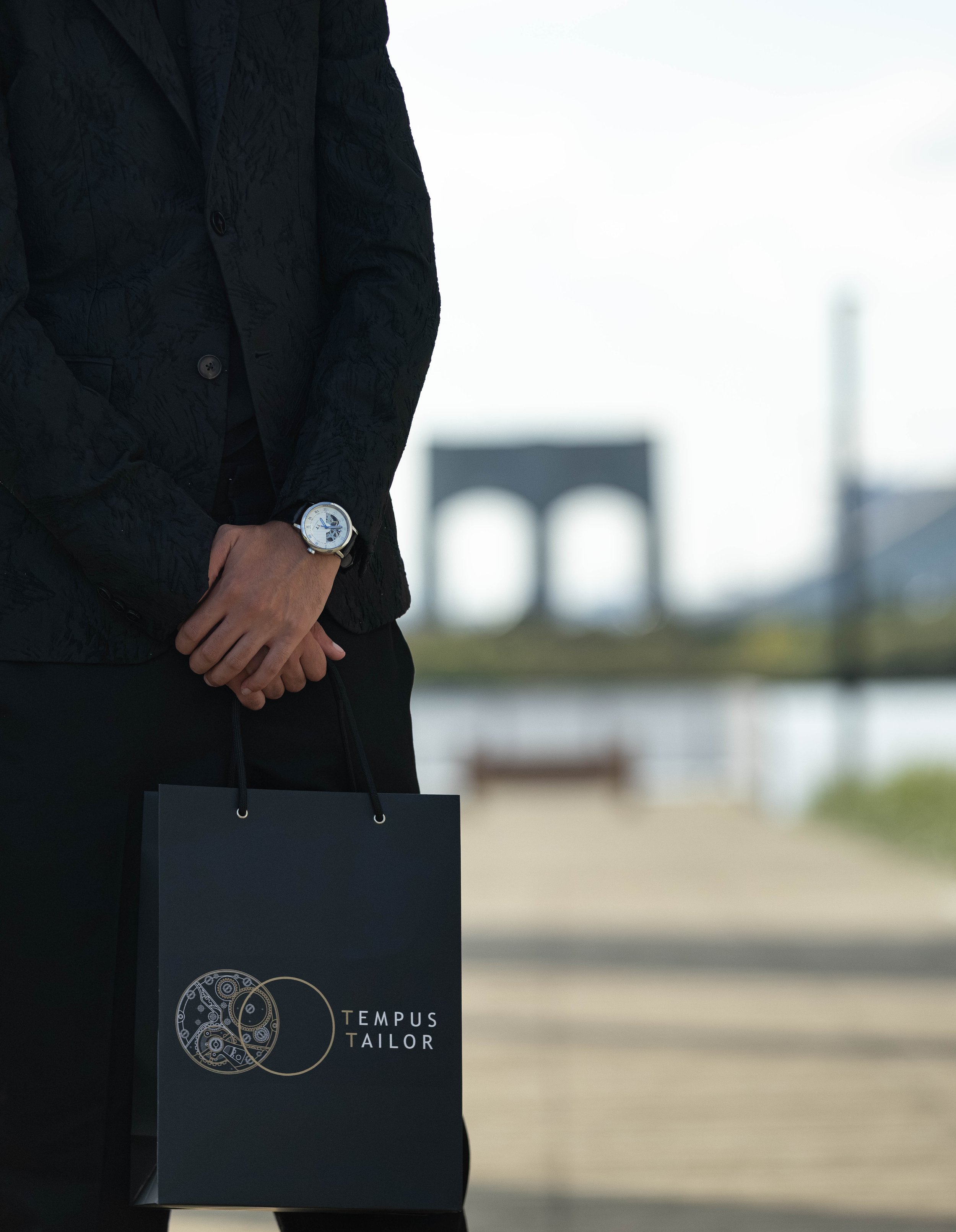 Close-up of a person wearing a black quilted jacket and a wristwatch, holding a black shopping bag with a logo that reads 'TEMPUS TAILOR'. The background features a blurred view of the Brooklyn Bridge.