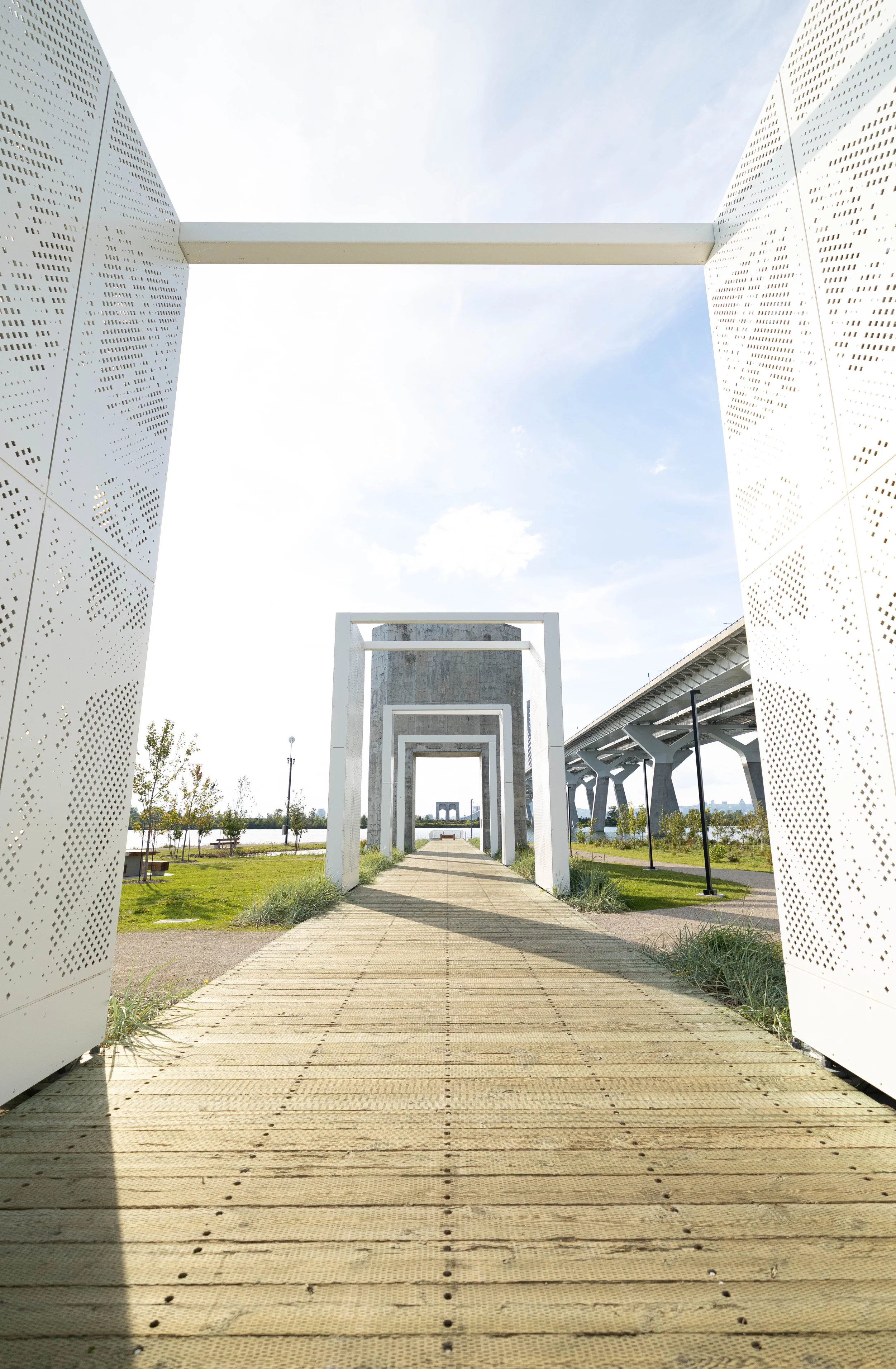 Photograph of a modern outdoor walkway with white framed arches and perforated metal panels on either side, leading toward a distant large, cubic structure, with grass and trees in the background under a partly cloudy sky.