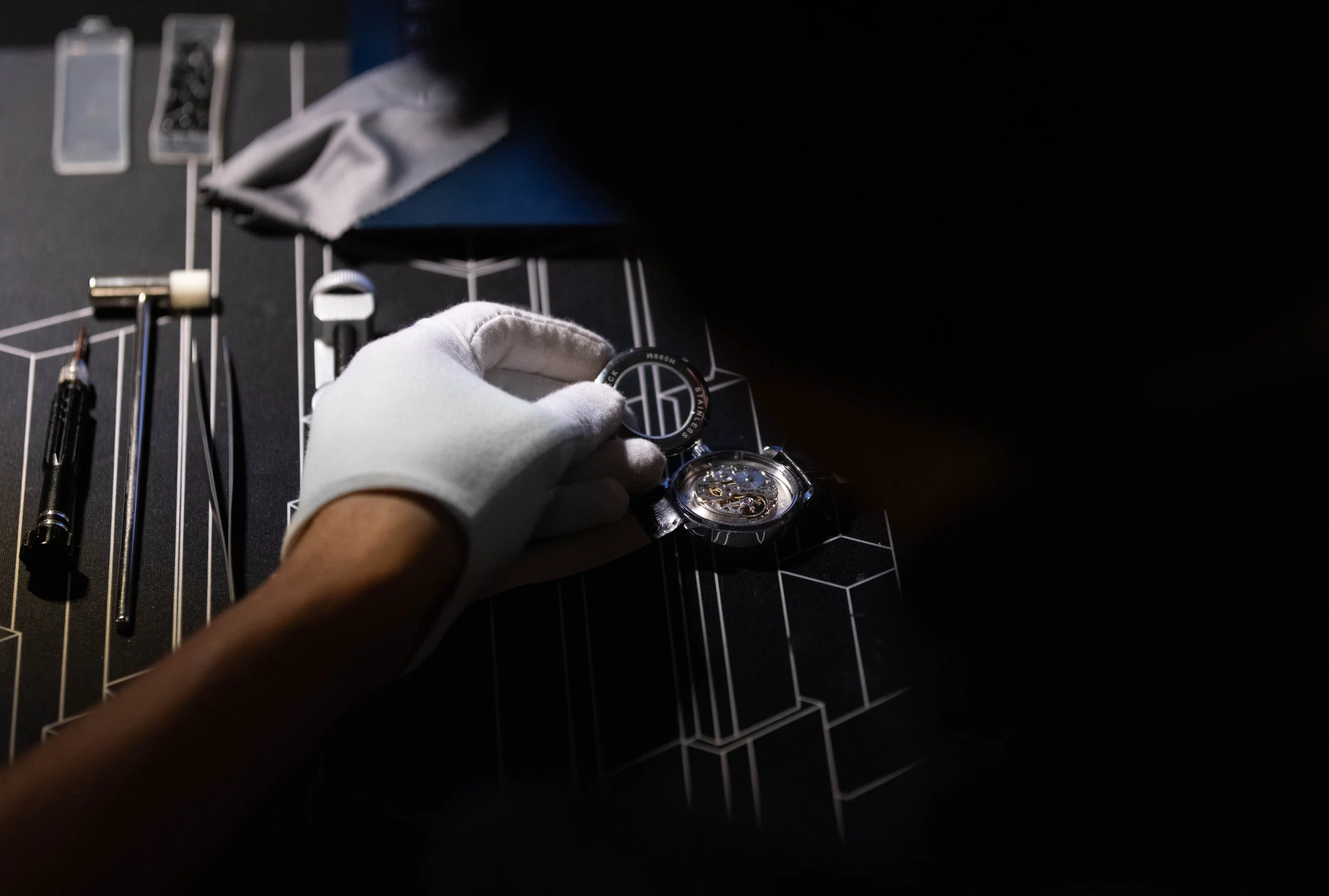 A person wearing a white glove holding a wristwatch over a black desk with watchmaking tools and a watch face.