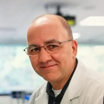 A man with glasses, bald on top with short hair on the sides, wearing a white lab coat, in what appears to be a medical or laboratory setting.