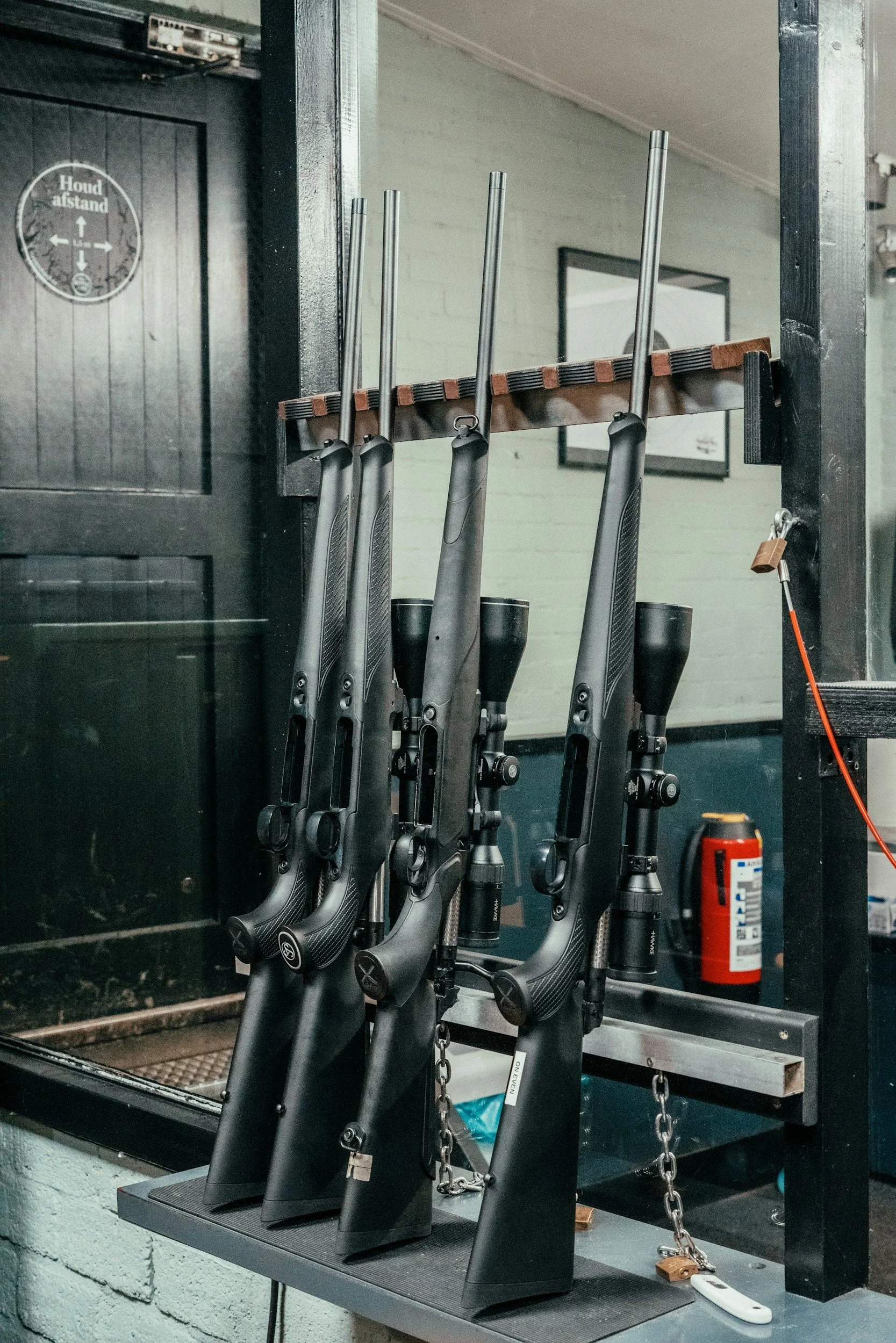 A wall-mounted rack holding four rifles with black stocks and metallic barrels, with safety chains attached to protect them. A fire extinguisher is placed below the rack, and a mirror and framed picture are on the wall behind.