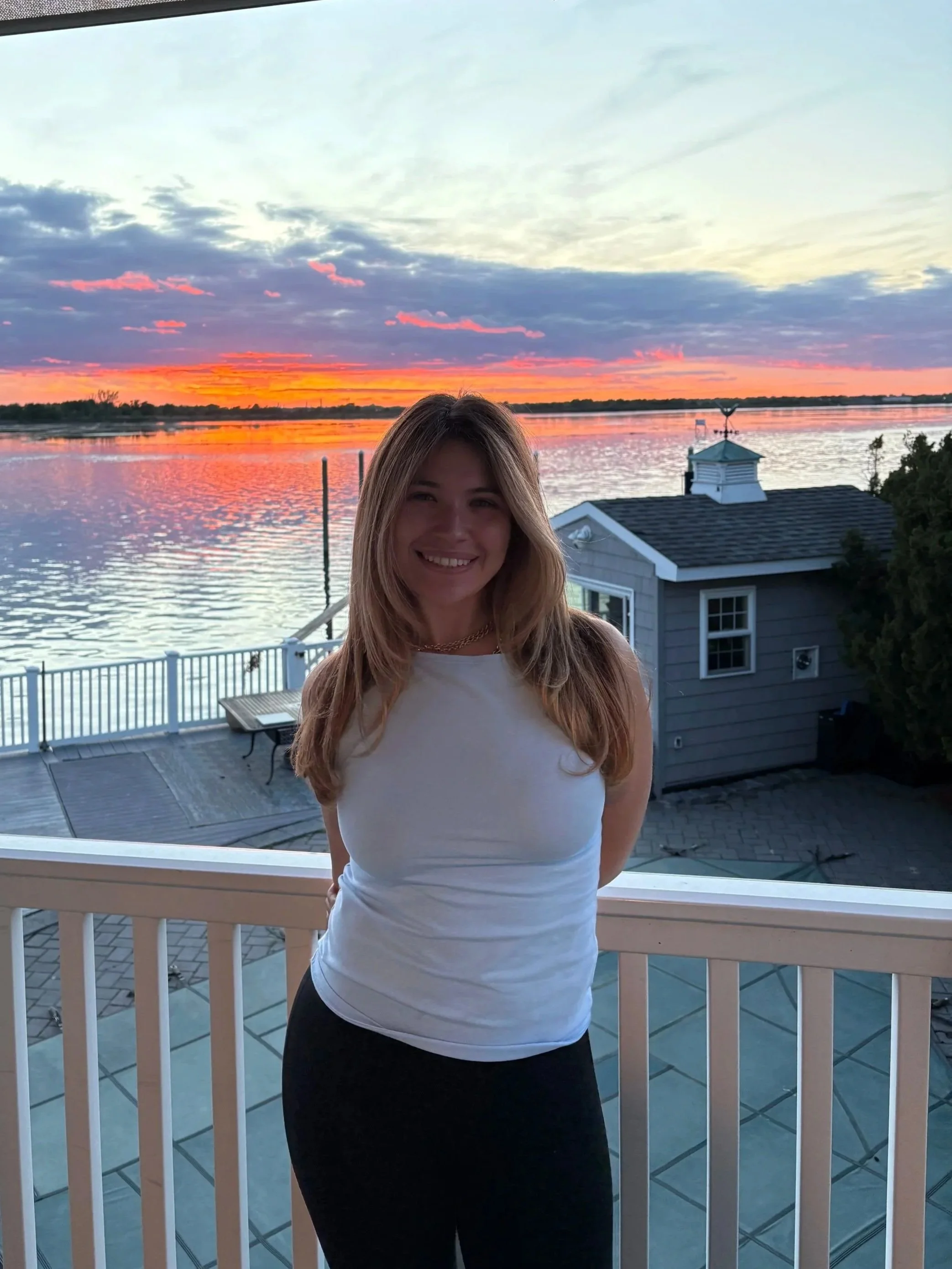 A woman standing on a balcony overlooking a body of water during sunset, with colorful clouds in the sky and a house with a gray roof in the background.
