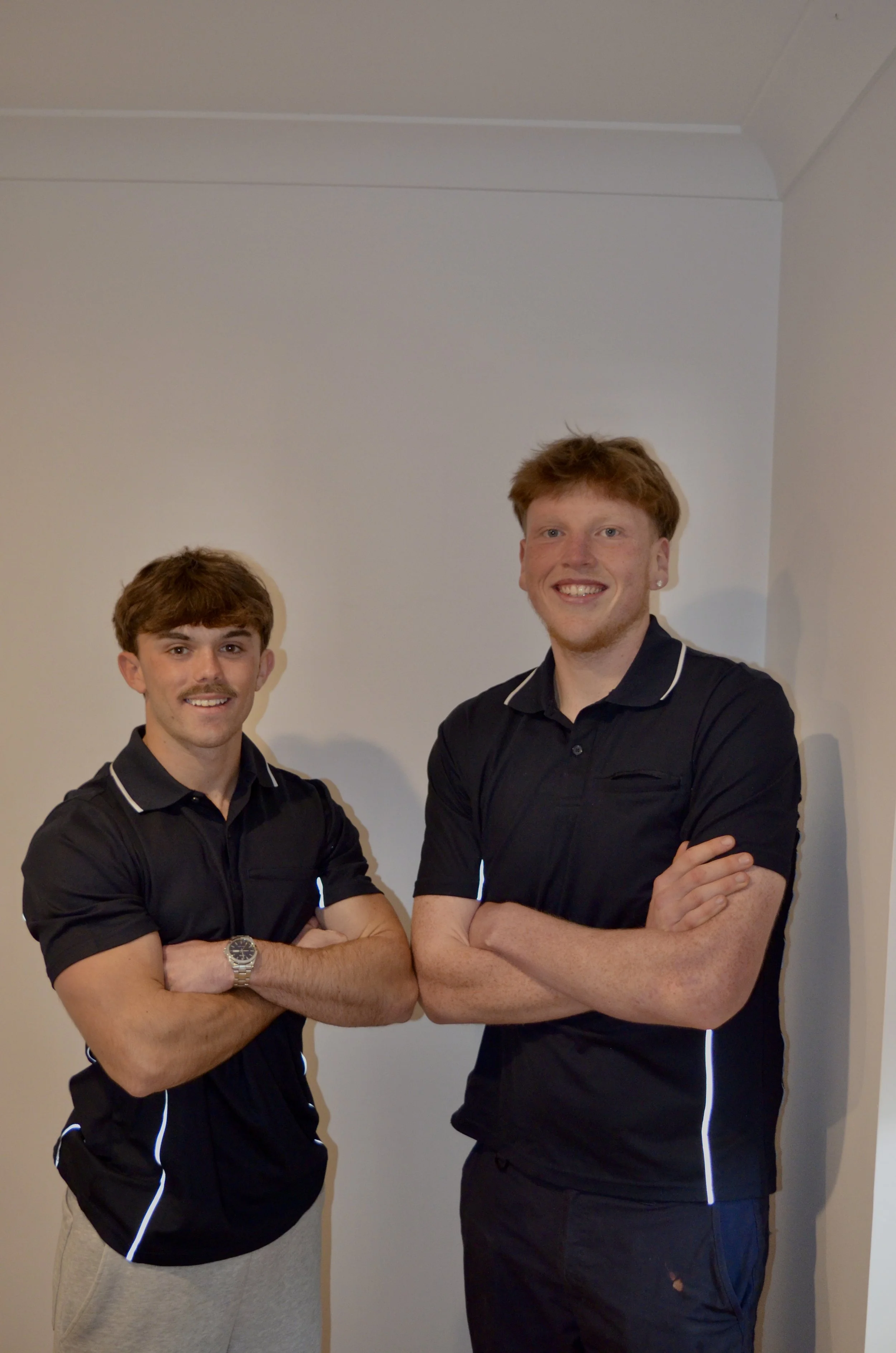 Two young men with red hair, wearing black polo shirts with white accents, standing with arms crossed, smiling at the camera against a plain wall.