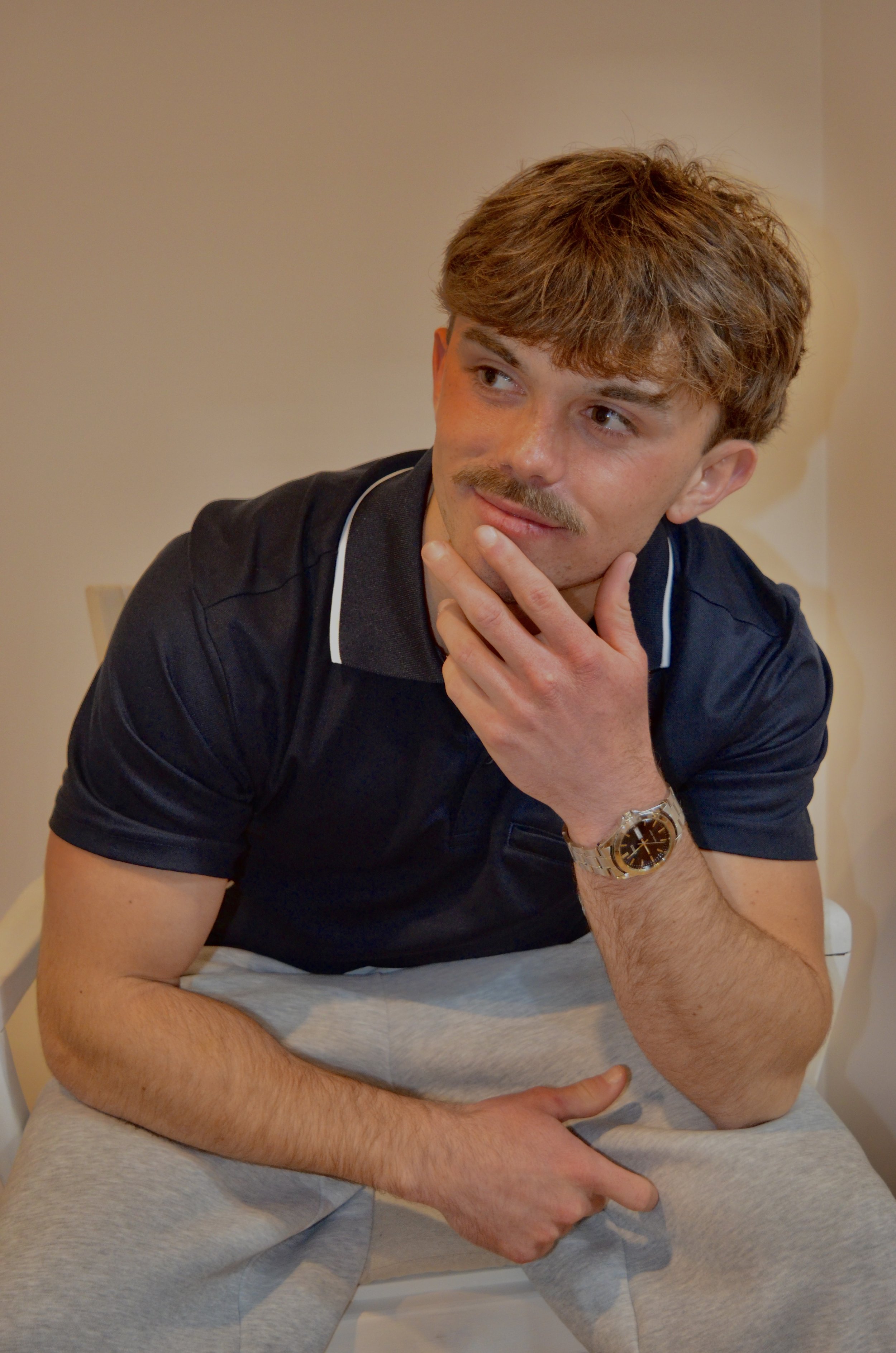 A young man with brown hair and a mustache, wearing a black polo shirt with white trim, sitting at a table with a beige wall background. He has a wristwatch on his left wrist and is resting his chin on his left hand, looking to the side.