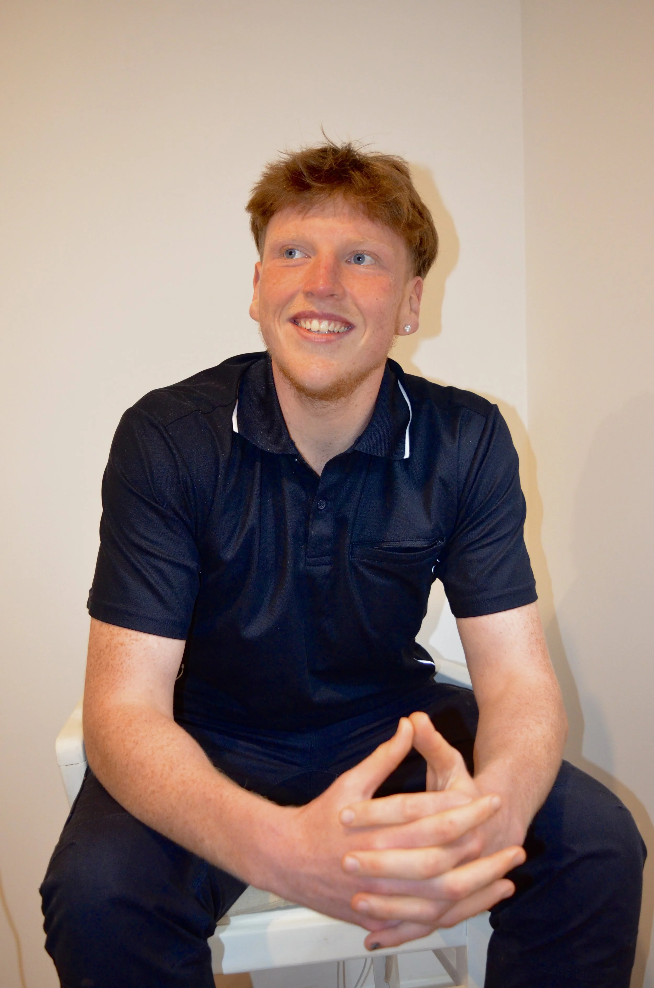 A young man with red hair and freckles, wearing a dark polo shirt, is sitting on a white chair against a plain beige wall, smiling and looking to his left.