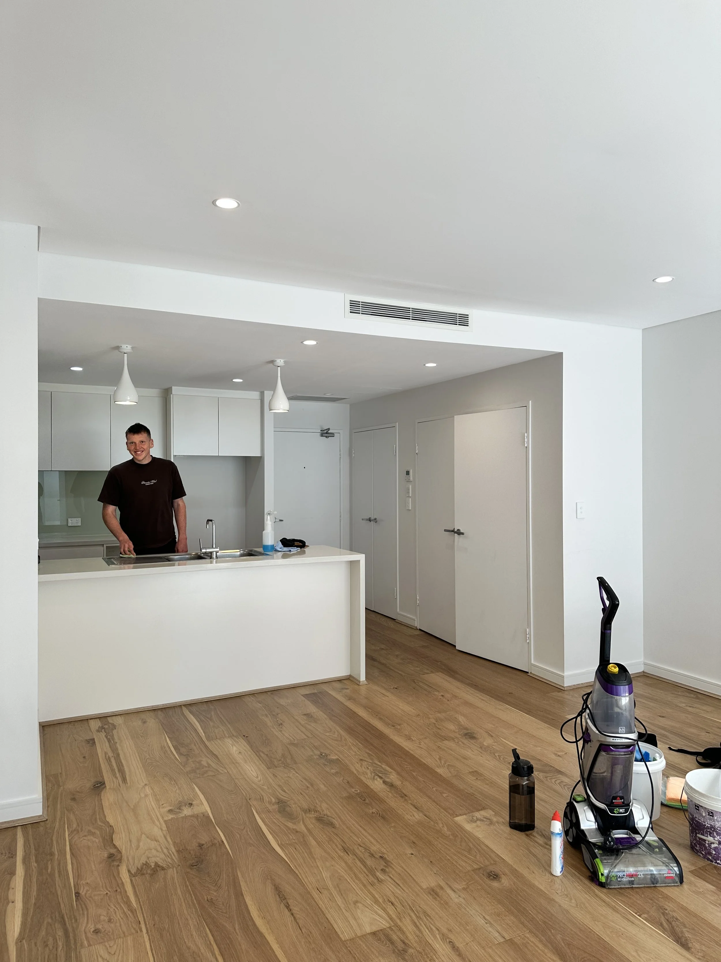 A man in a brown shirt standing in a modern kitchen with white cabinets and wood floors, and cleaning equipment on the floor.