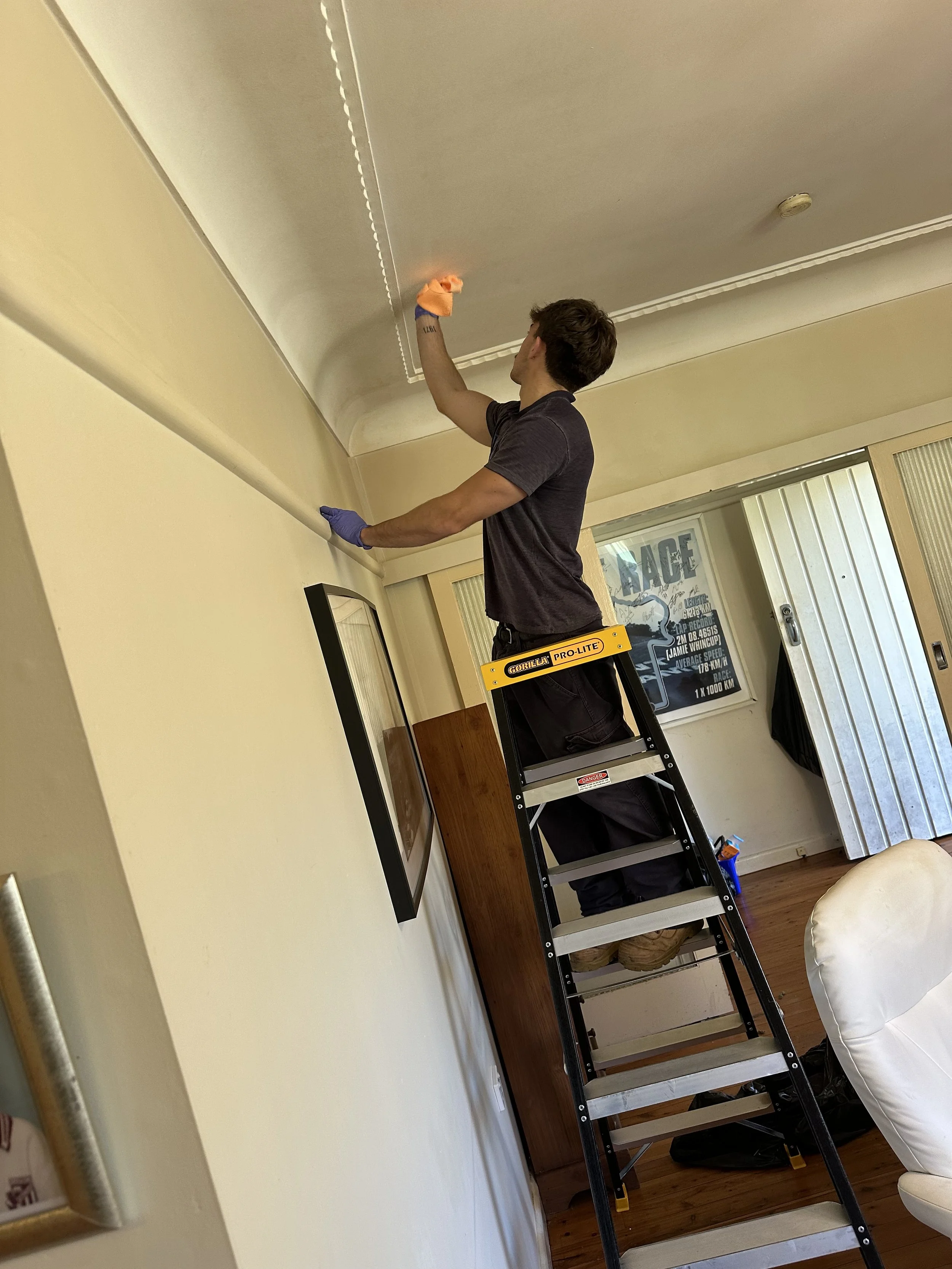 A person stands on a ladder and is cleaning or working on the ceiling in a room. The person is wearing a black shirt, black pants, gloves, and tan work boots. The room has beige walls, a window with blinds, and a framed picture on the wall.