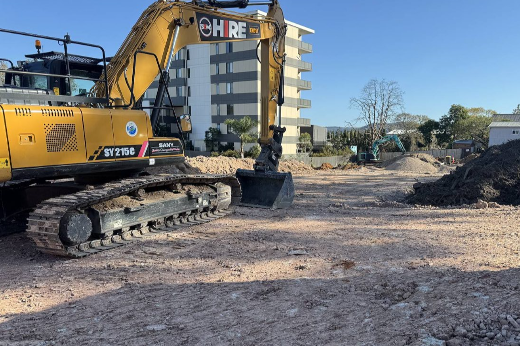 A yellow excavator on a construction site, with piles of dirt and gravel around. There is a tall building and some trees in the background, under a clear blue sky.