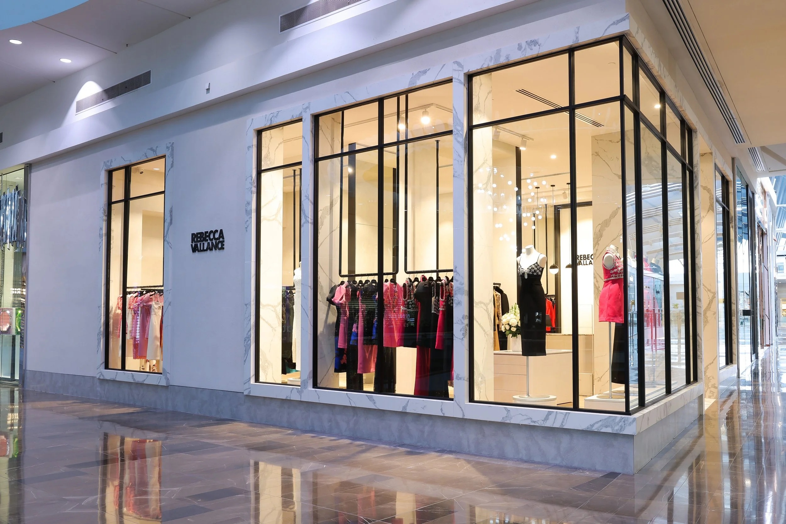 Clothing store named Rebecca Vallance with large display windows showcasing dresses and mannequins inside. The exterior features a marble-like facade and modern lighting.