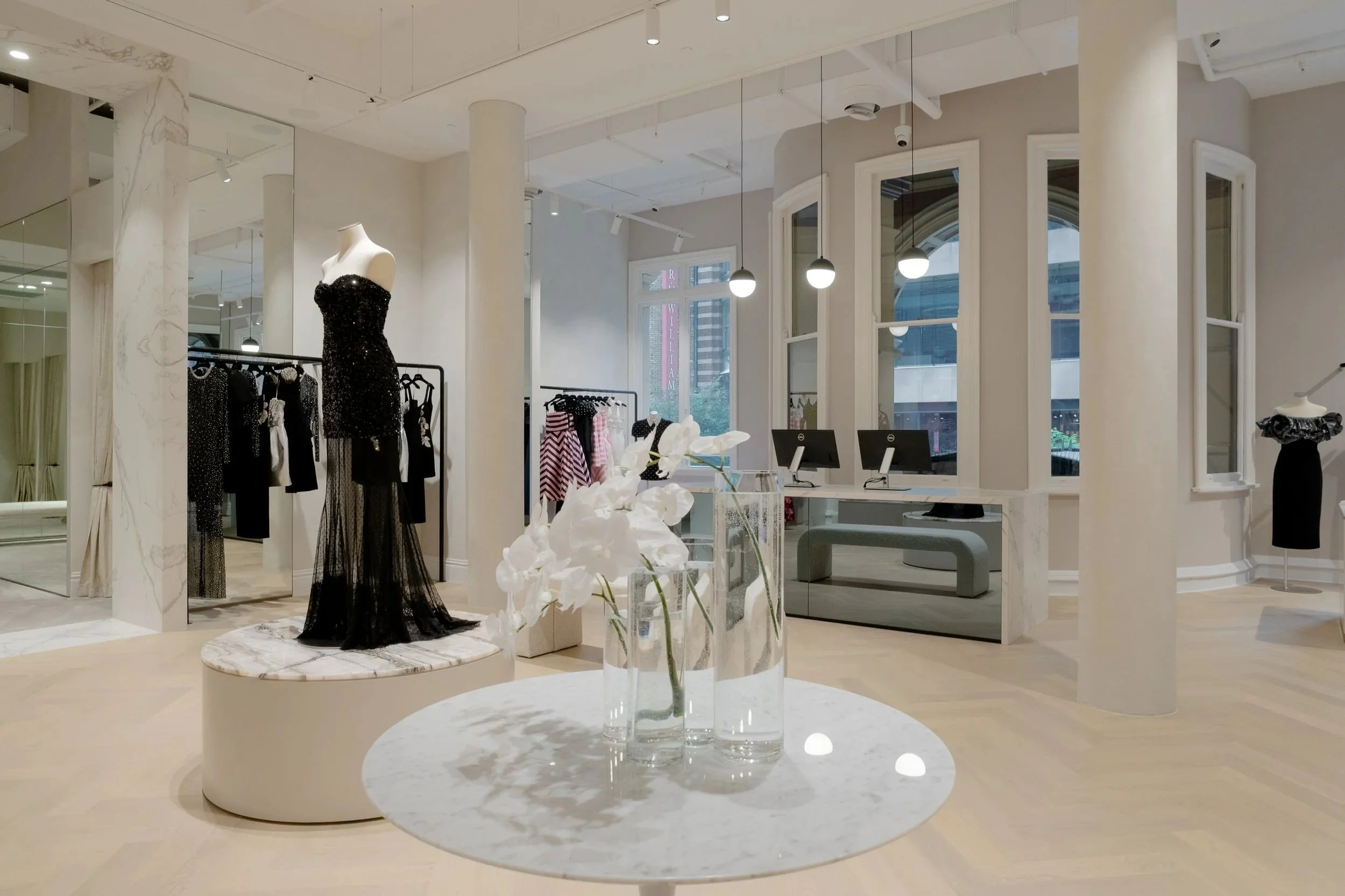 Inside a modern clothing store displaying elegant black dresses on mannequins and clothing racks, with a white marble table holding flowers and fitting counters with computer monitors near large windows.