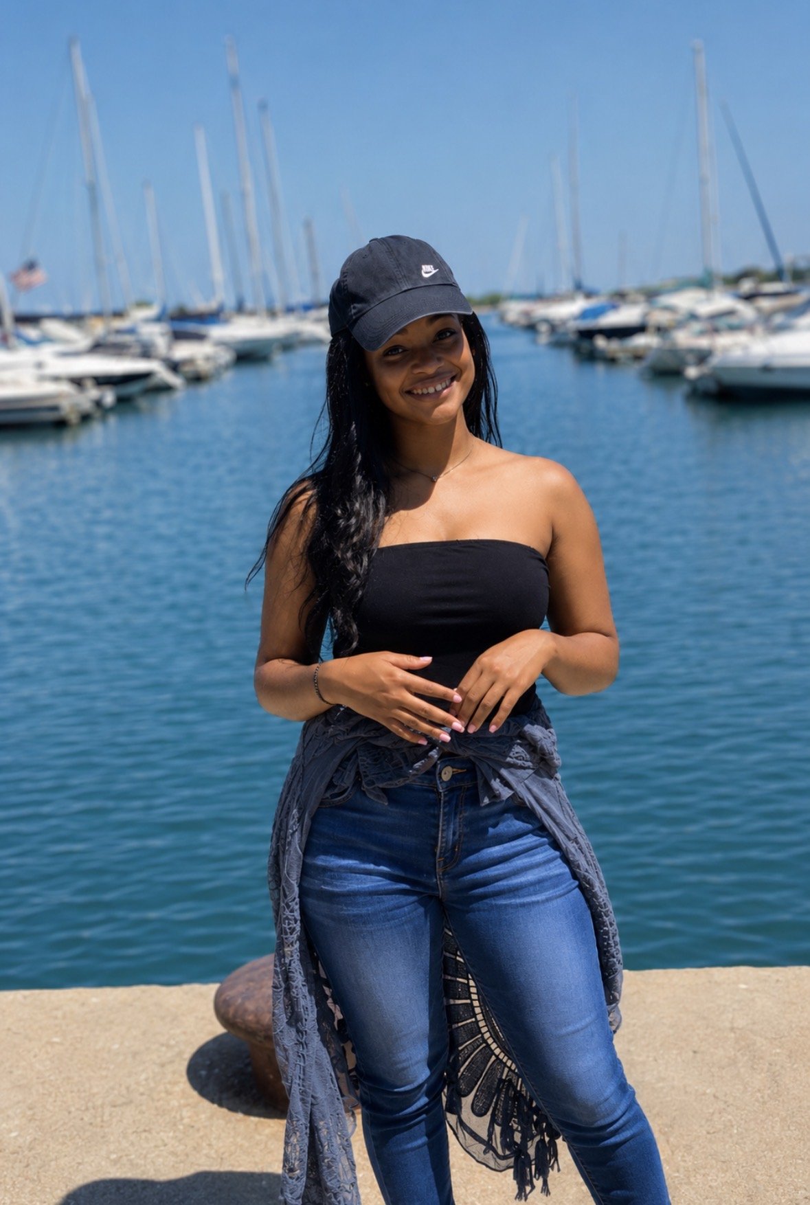 A woman with long black hair wearing a black strapless top, blue jeans, a black Nike cap, and a gray patterned jacket tied around her waist, standing in front of a marina with numerous sailboats and yachts on a sunny day.