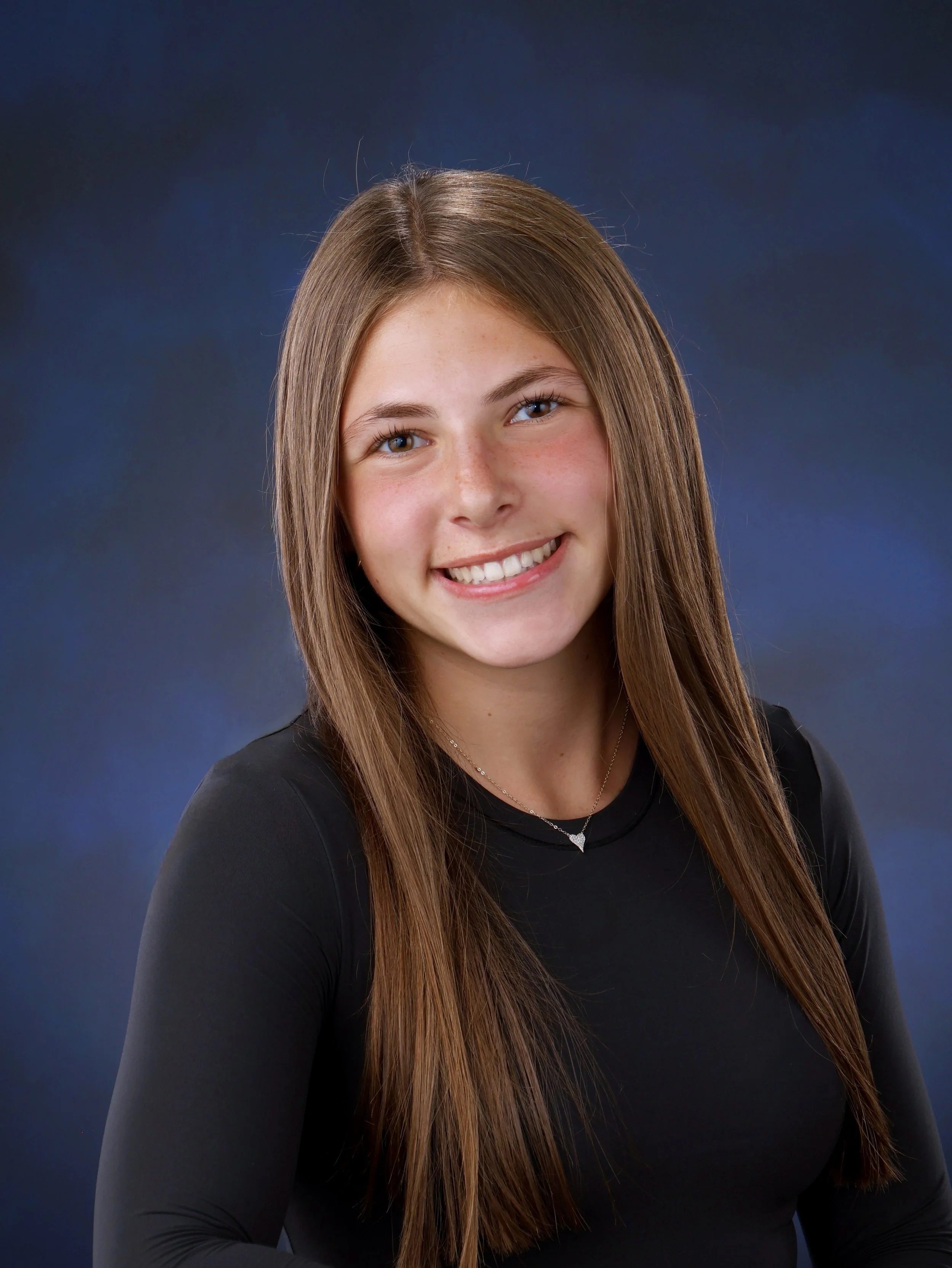 A young woman with long brown hair smiling, wearing a black top and a necklace, against a dark blue background.
