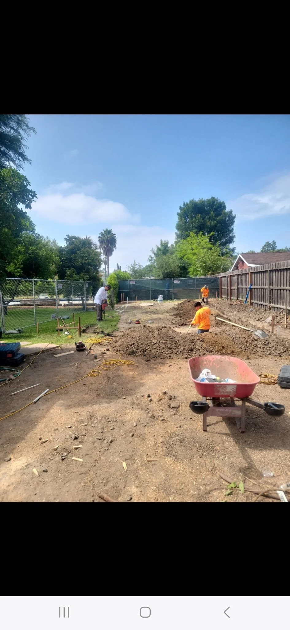 Construction workers digging and leveling soil in a backyard, with tools, a wheelbarrow, and fencing around the work area.