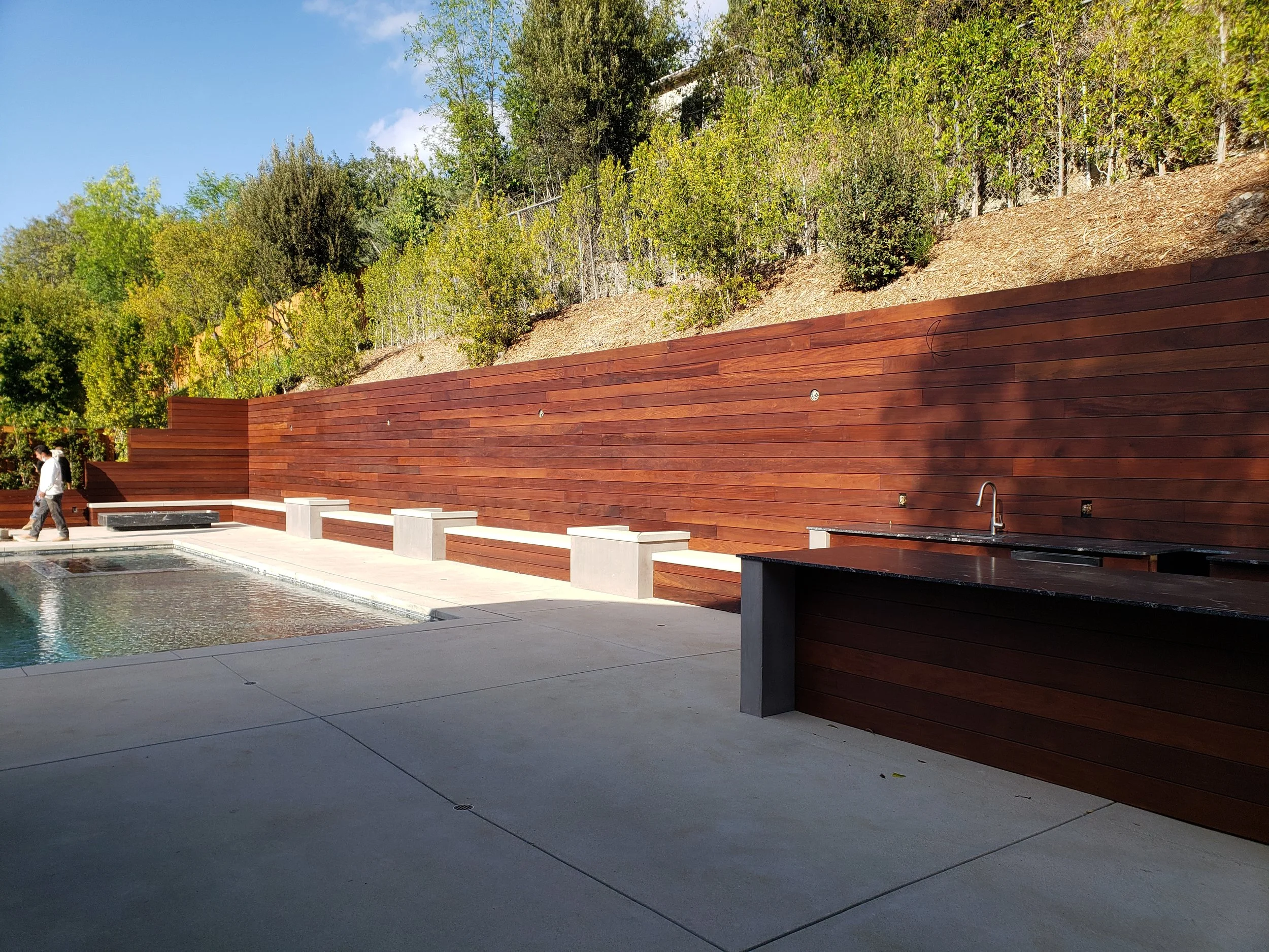 An outdoor area with a swimming pool, a wooden privacy wall, and trees in the background under a clear blue sky.